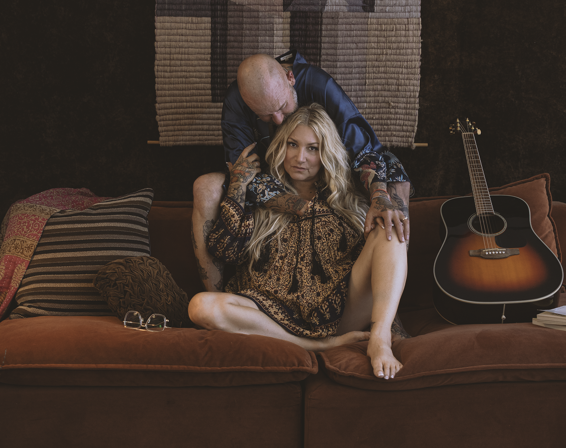 A woman with long blonde hair and tattoos sitting on a brown couch, with a man with a shaved head leaning over her from behind. There is an acoustic guitar on the right side of the couch.