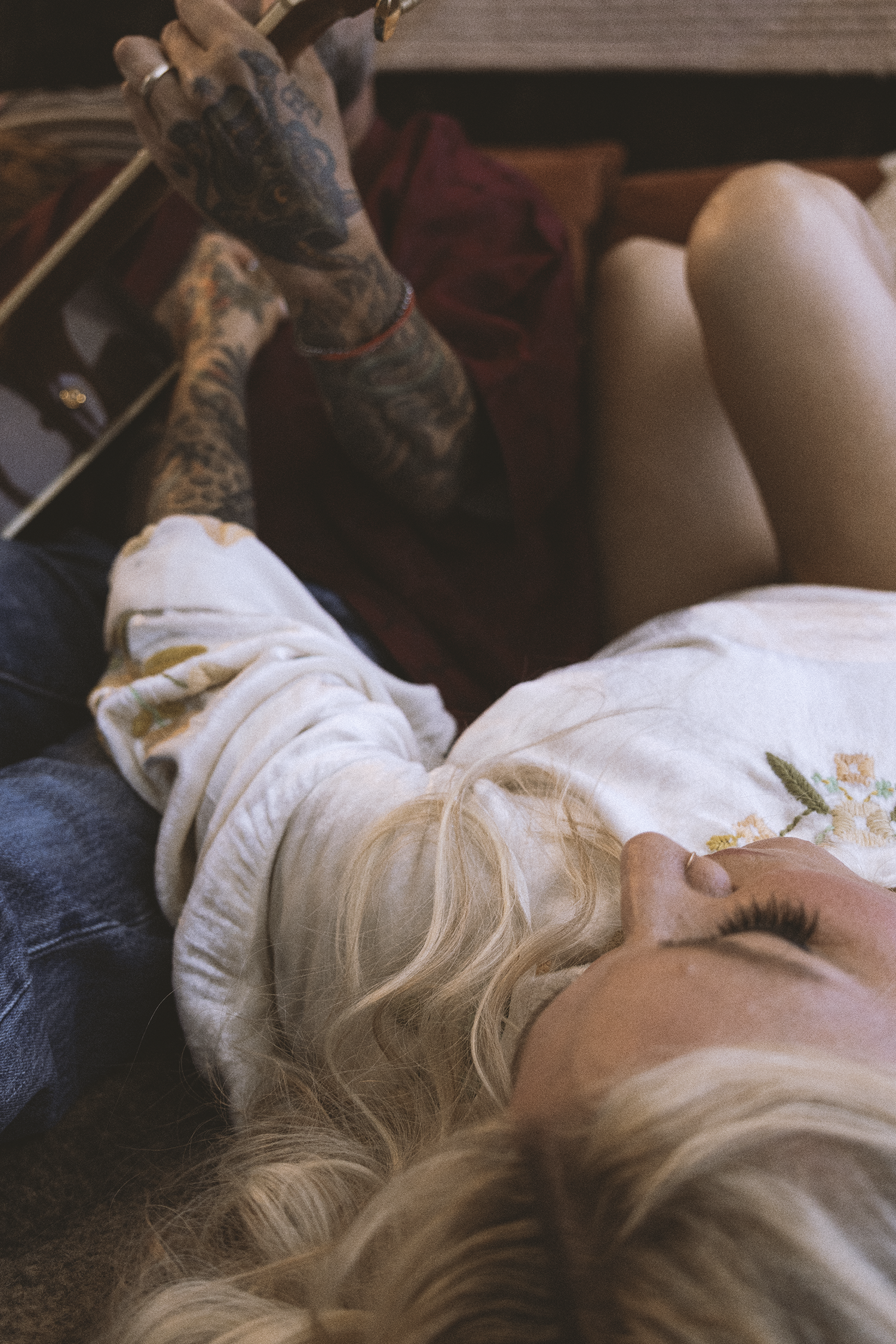 A woman with long blonde hair lying on a bed, wearing a white embroidered top. A person with heavily tattooed arms playing a guitar nearby.