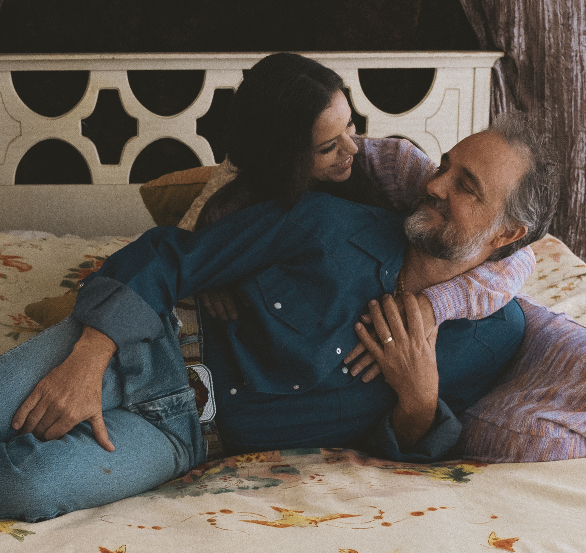 A woman and man are lying on a bed, smiling and embracing each other warmly. The man is wearing vintage Wrangler denim shirt and jeans. 