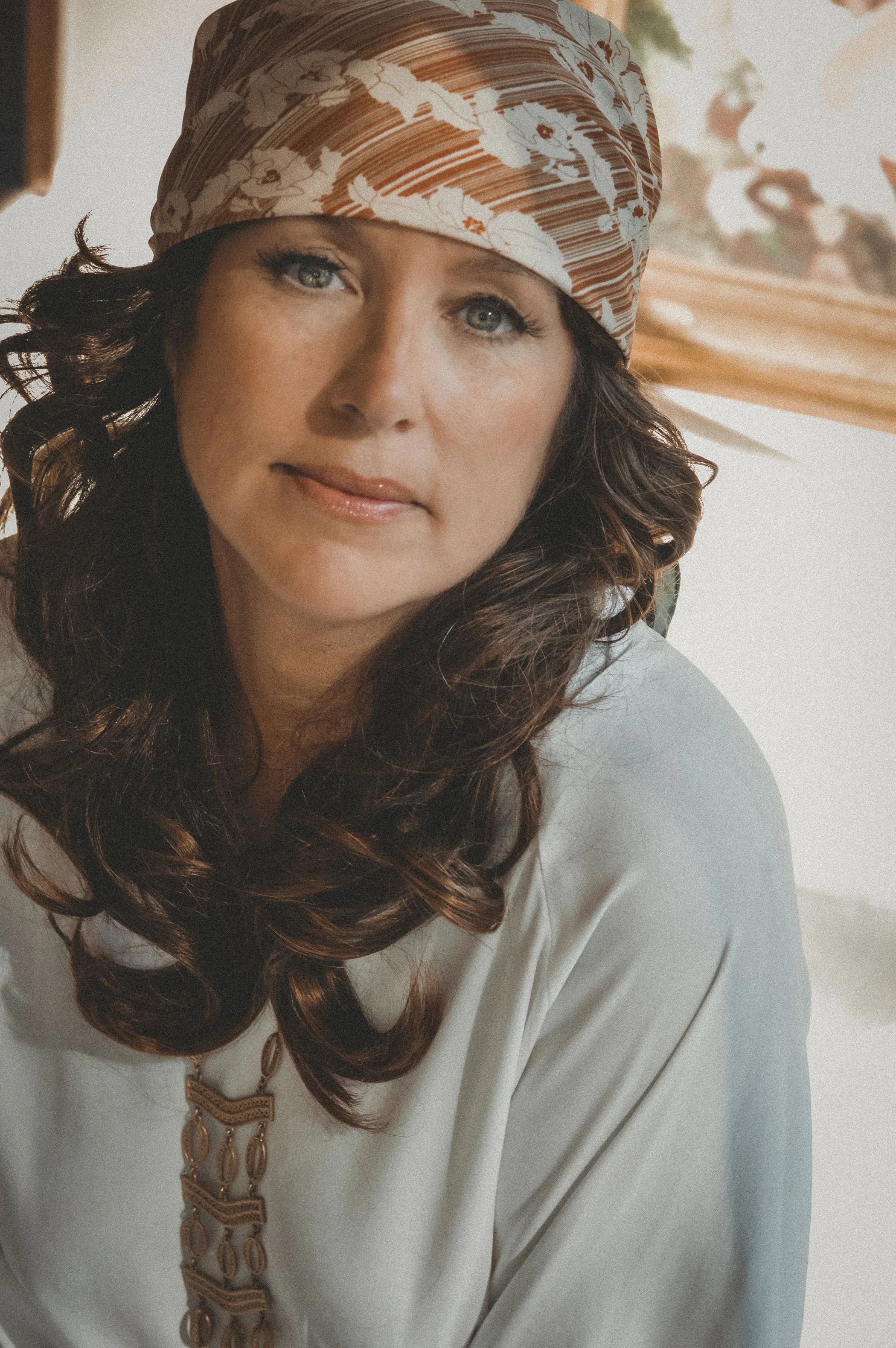 A woman with long curly brown hair wearing a patterned headscarf and a white blouse, looking at the camera, in a cozy indoor setting.