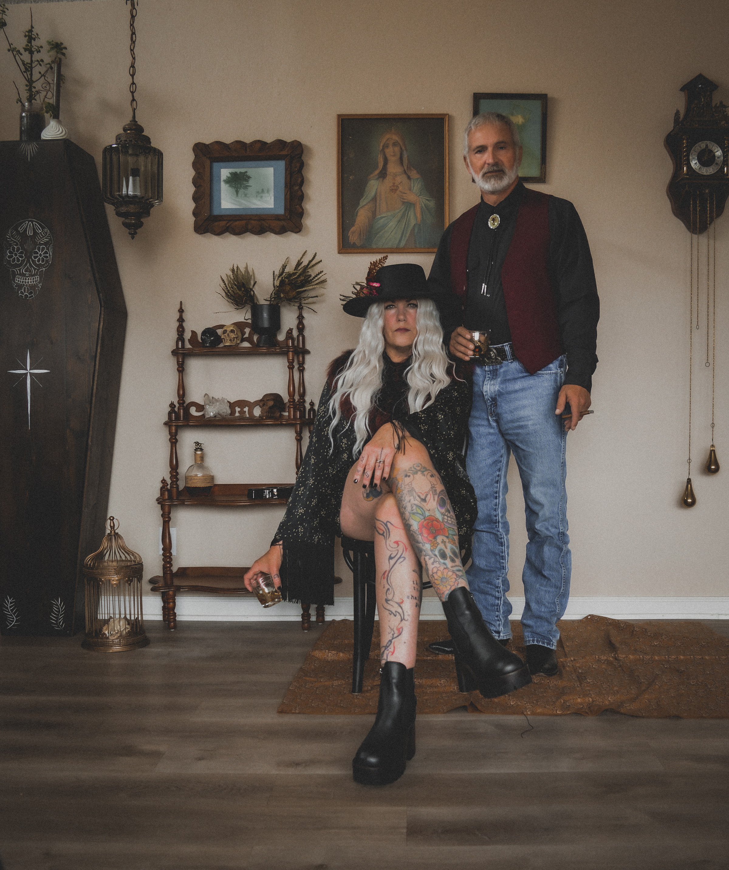 A woman with long white curly hair, tattoos, and black platform boots sits on a chair, holding a drink. A man with gray hair and beard, wearing a black shirt, red vest, and jeans, stands behind her holding a drink. They are in a decorated room with f