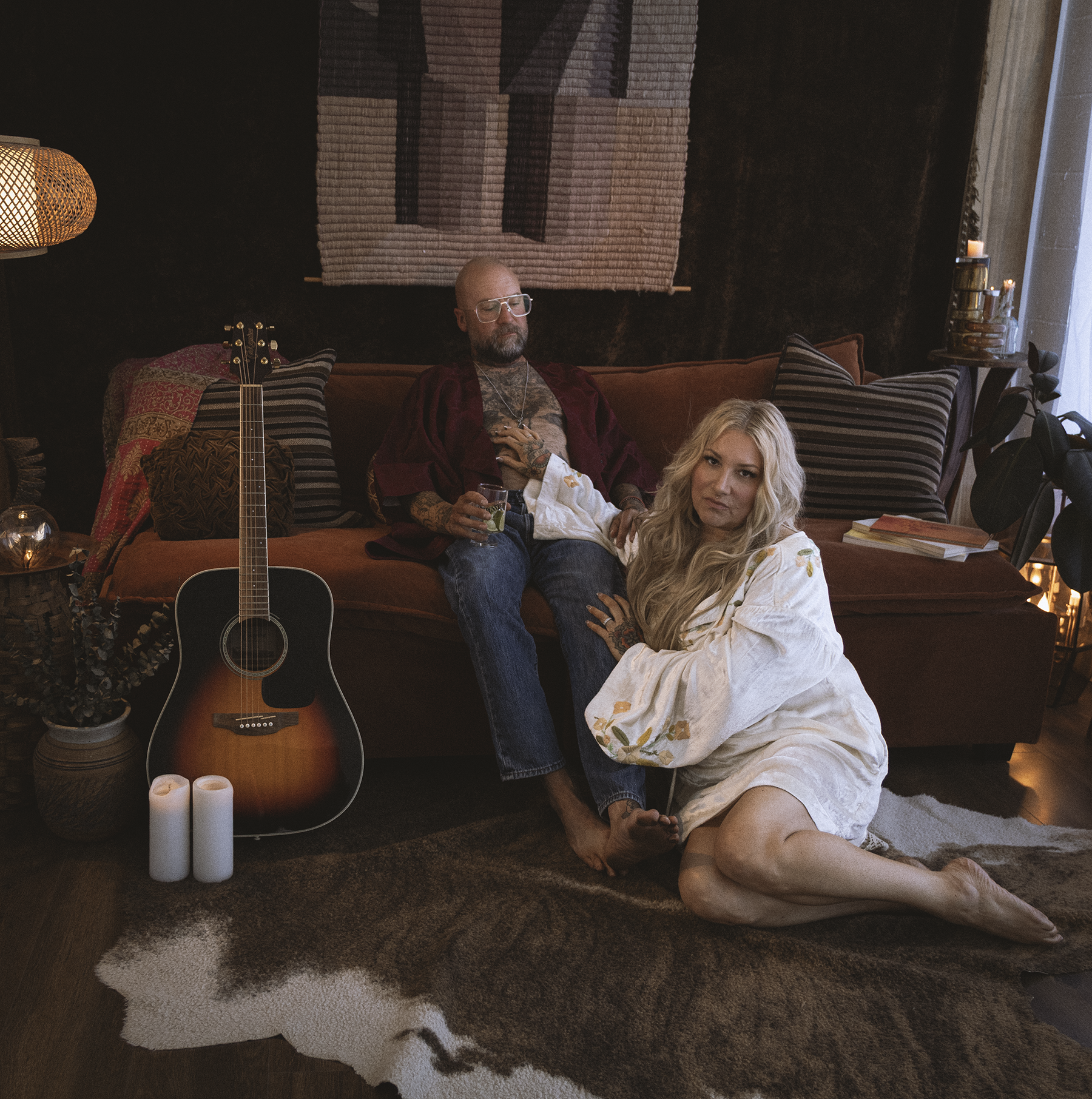 A man with tattoos and glasses relaxing on a sofa, holding a drink, with a woman in a white dress sitting on the floor beside him, in a dimly lit cozy living room with a guitar, candles, and decor.