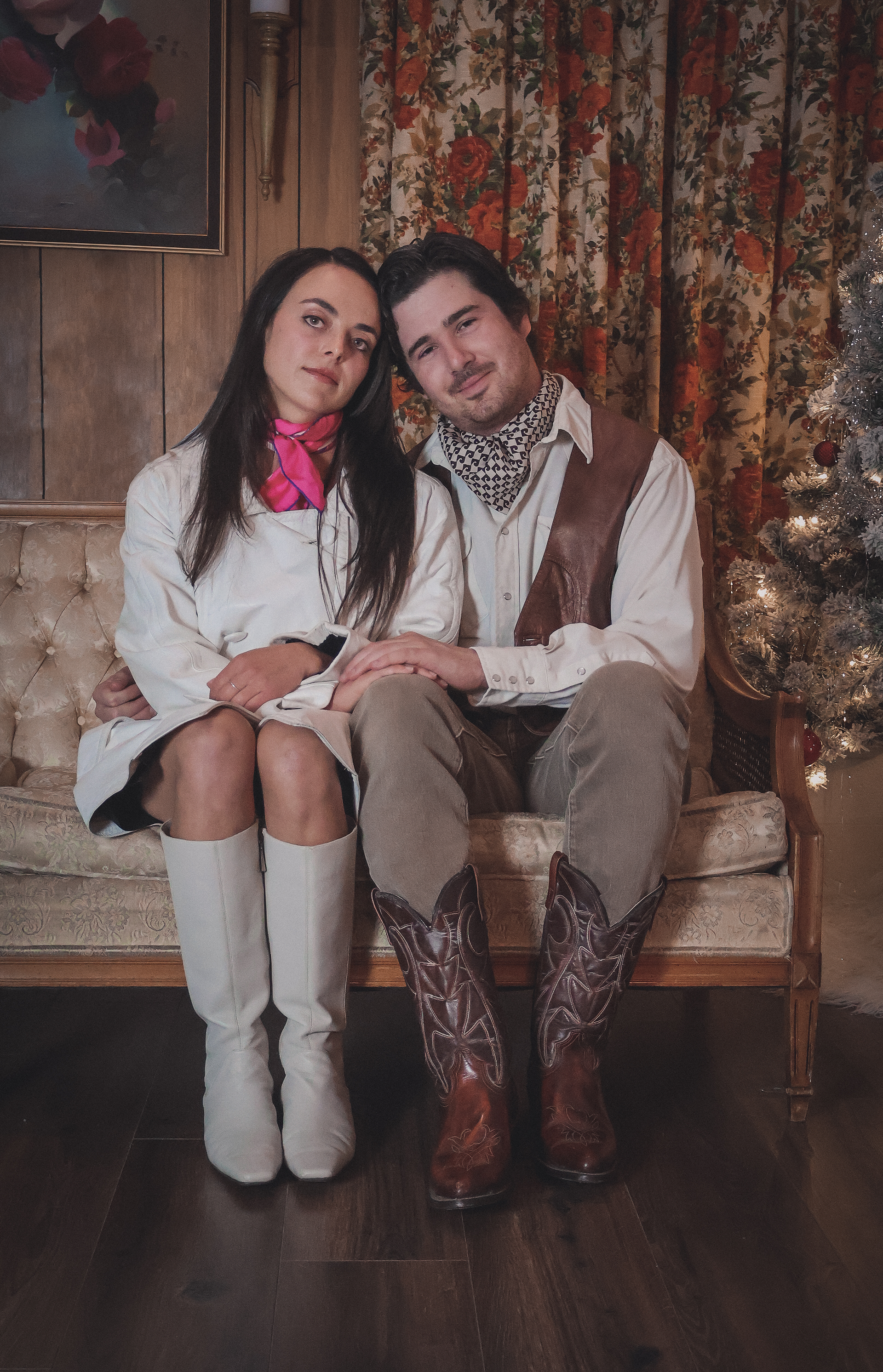 A young woman and young man sitting close together on a vintage sofa in a cozy, rustic room decorated for Christmas, with a Christmas tree on the right and floral curtains in the background. The woman is wearing white knee-high boots, a white outfit,