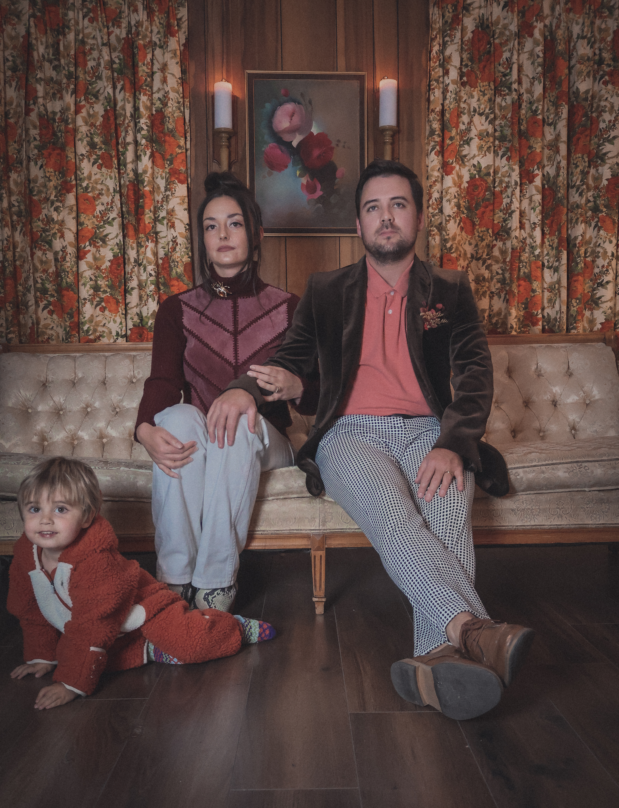 Three people sitting and kneeling on a vintage sofa in a room with floral curtains. The woman with dark hair and a high bun, wearing a maroon and purple top, sits with her arm on a man's knee. The man has dark hair and a beard, wearing a brown velvet