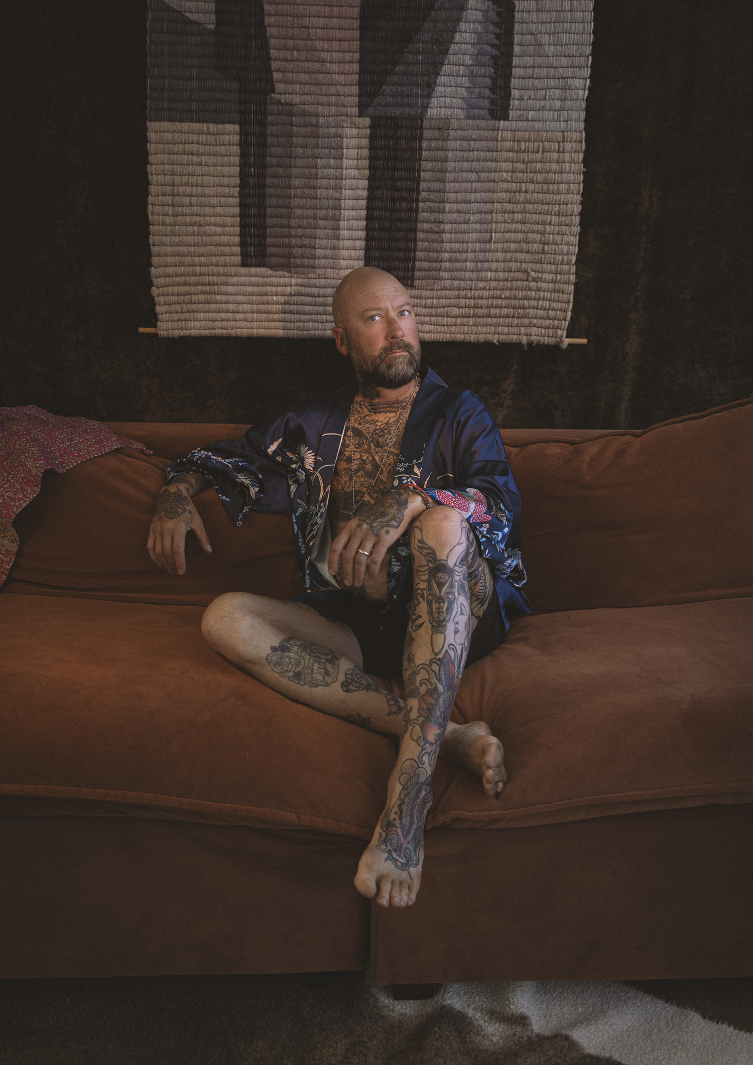 A tattooed man with a beard sitting on a brown couch, wearing a blue satin robe, in front of a textured wall hanging.
