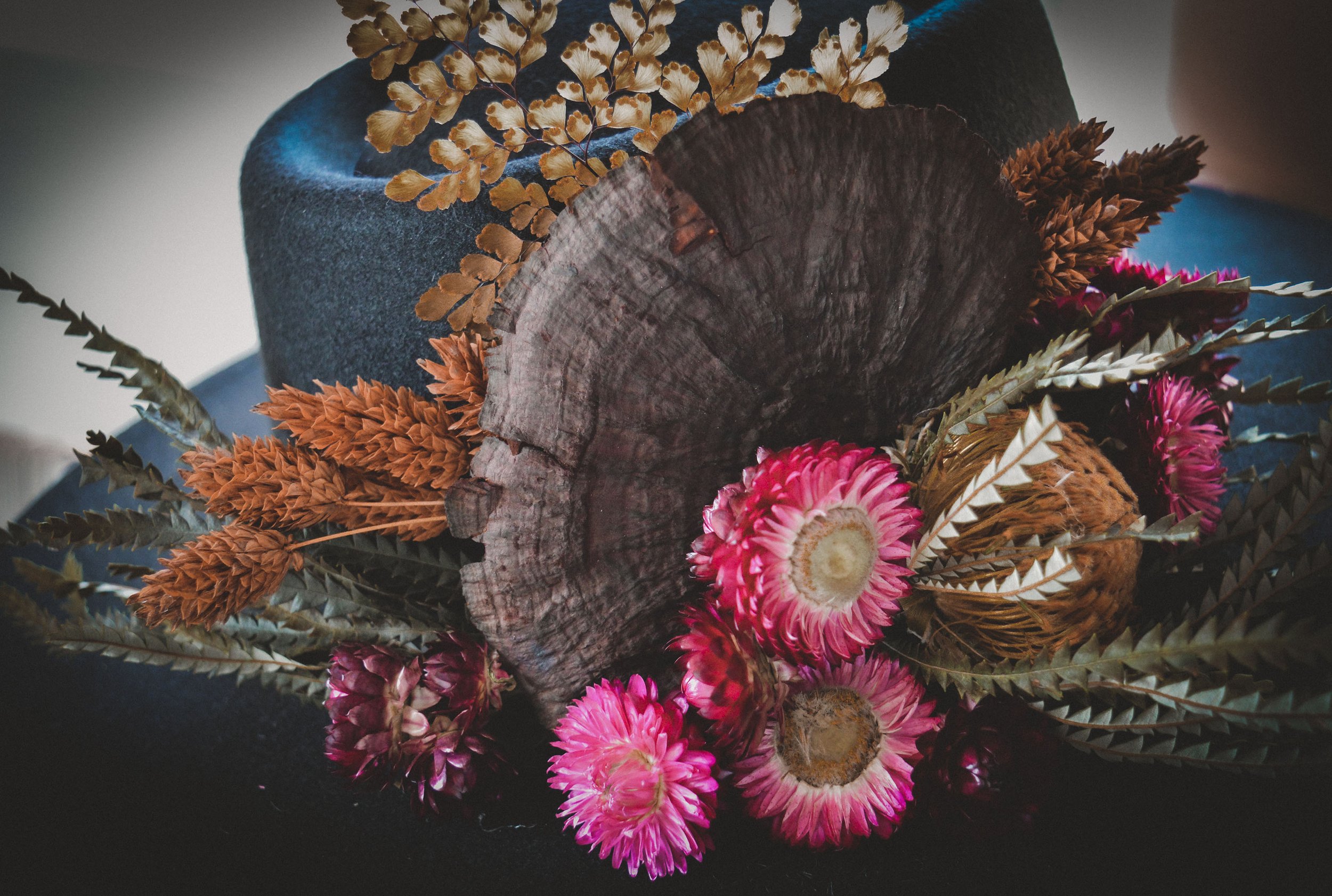 A rustic floral arrangement with pink flowers, dried grasses, and a large dried mushroom adorn a black felt hat.