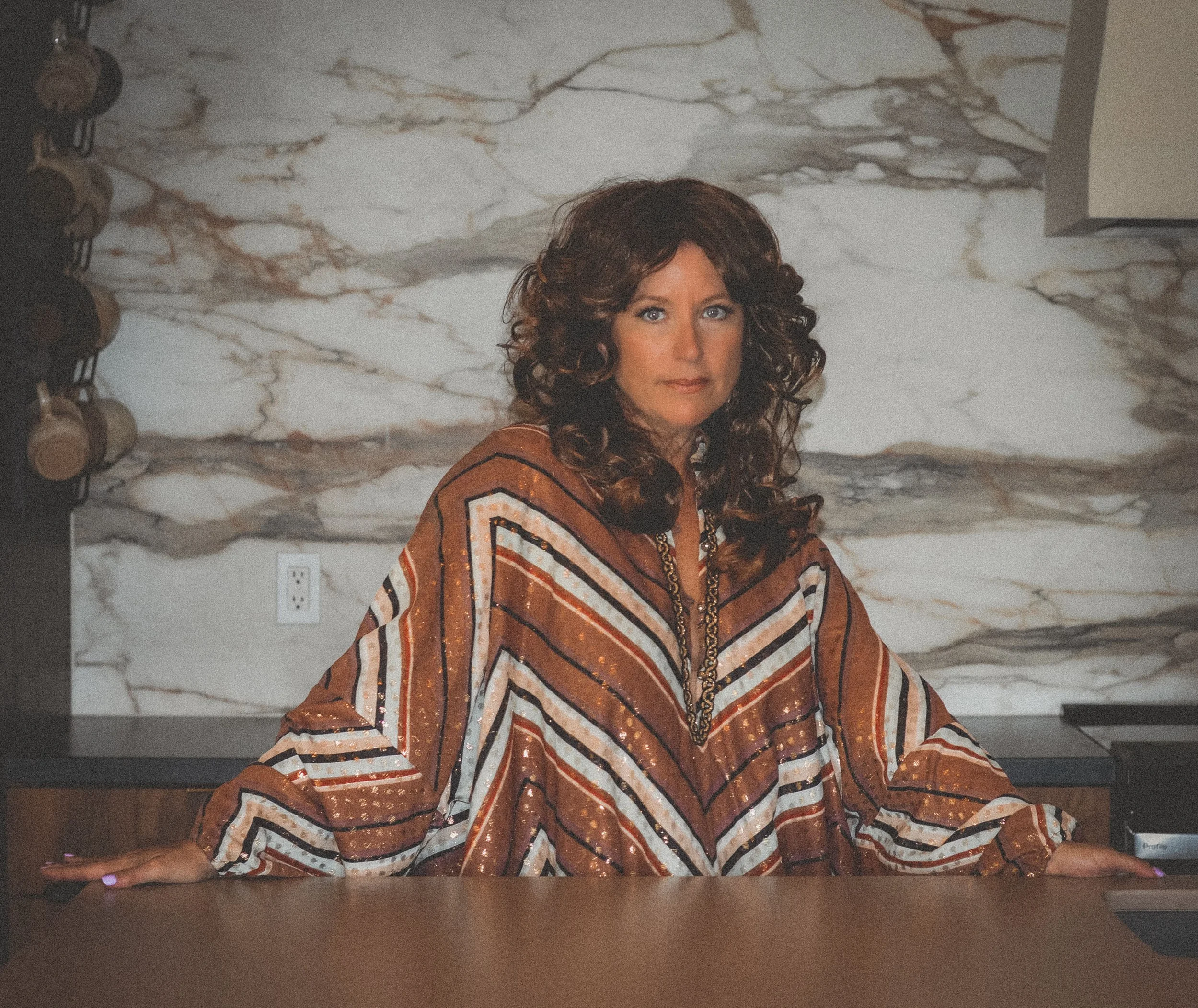 A woman with curly dark hair wearing a brown, white, and black vintage 70's striped caftan standing in a kitchen with a marble backsplash and a wooden countertop.