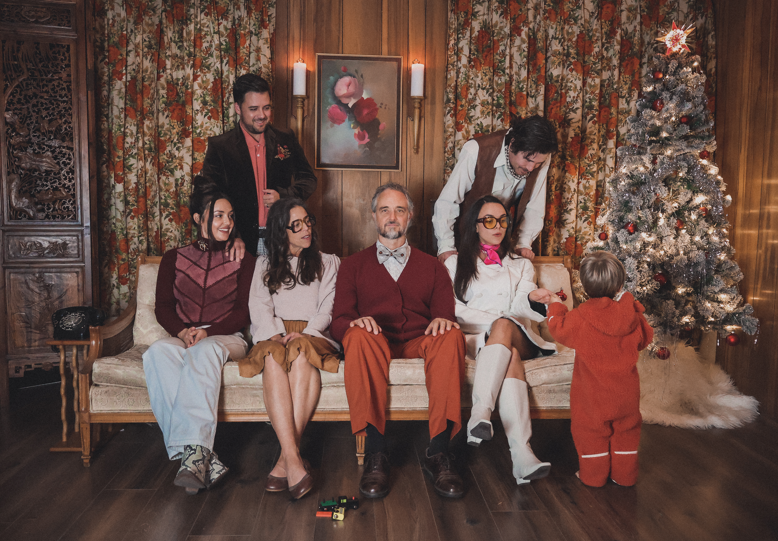 A group of six adults and one small child in a holiday scene. Four adults are sitting on a vintage sofa, two adults are standing behind the sofa and leaning forward. A small child dressed in red is facing the Christmas tree. The setting has wooden-pa