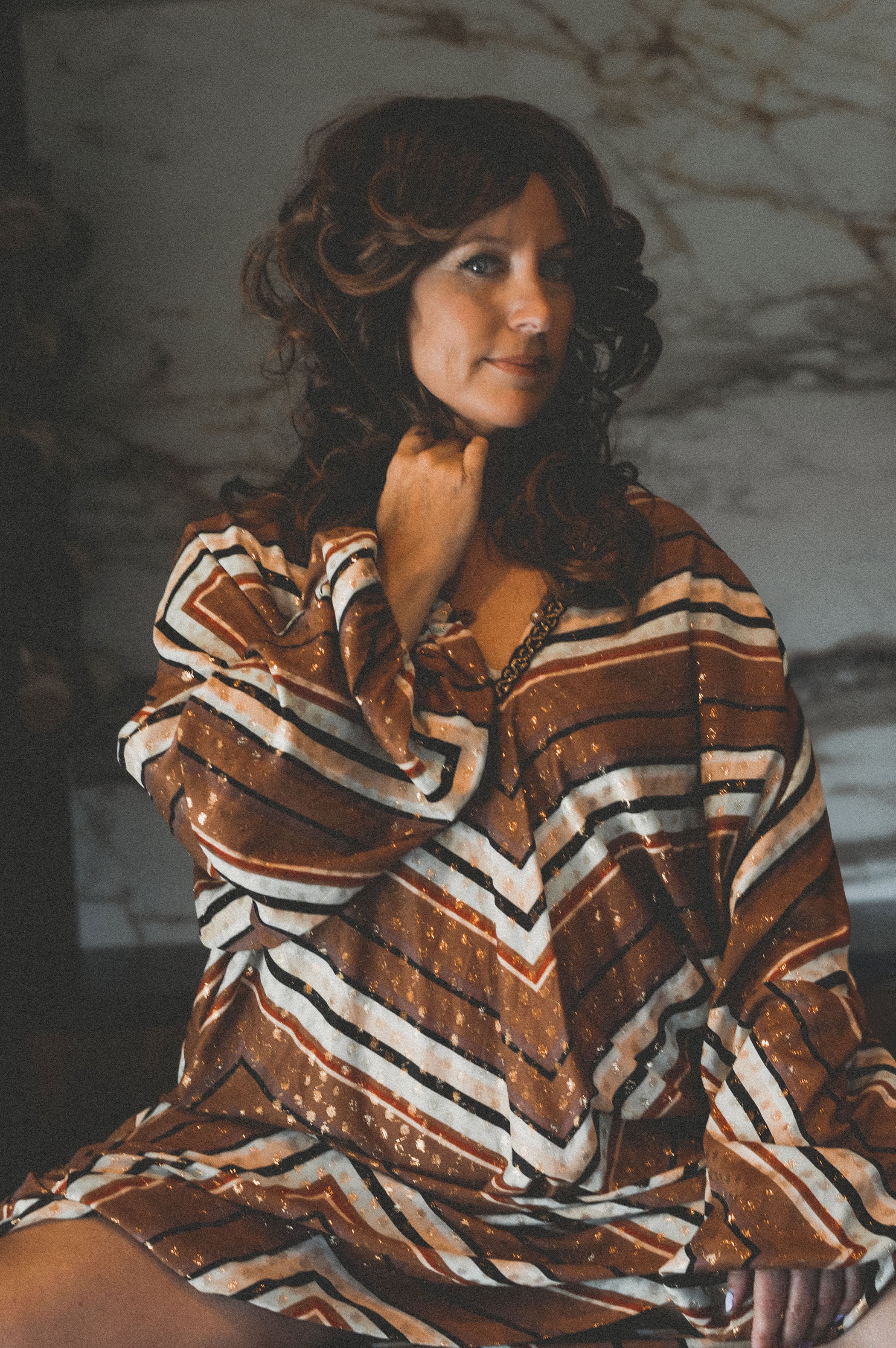 A woman with curly dark hair wearing a brown, white, and black vintage 70's  striped caftan sitting indoors in front of a marble wall.