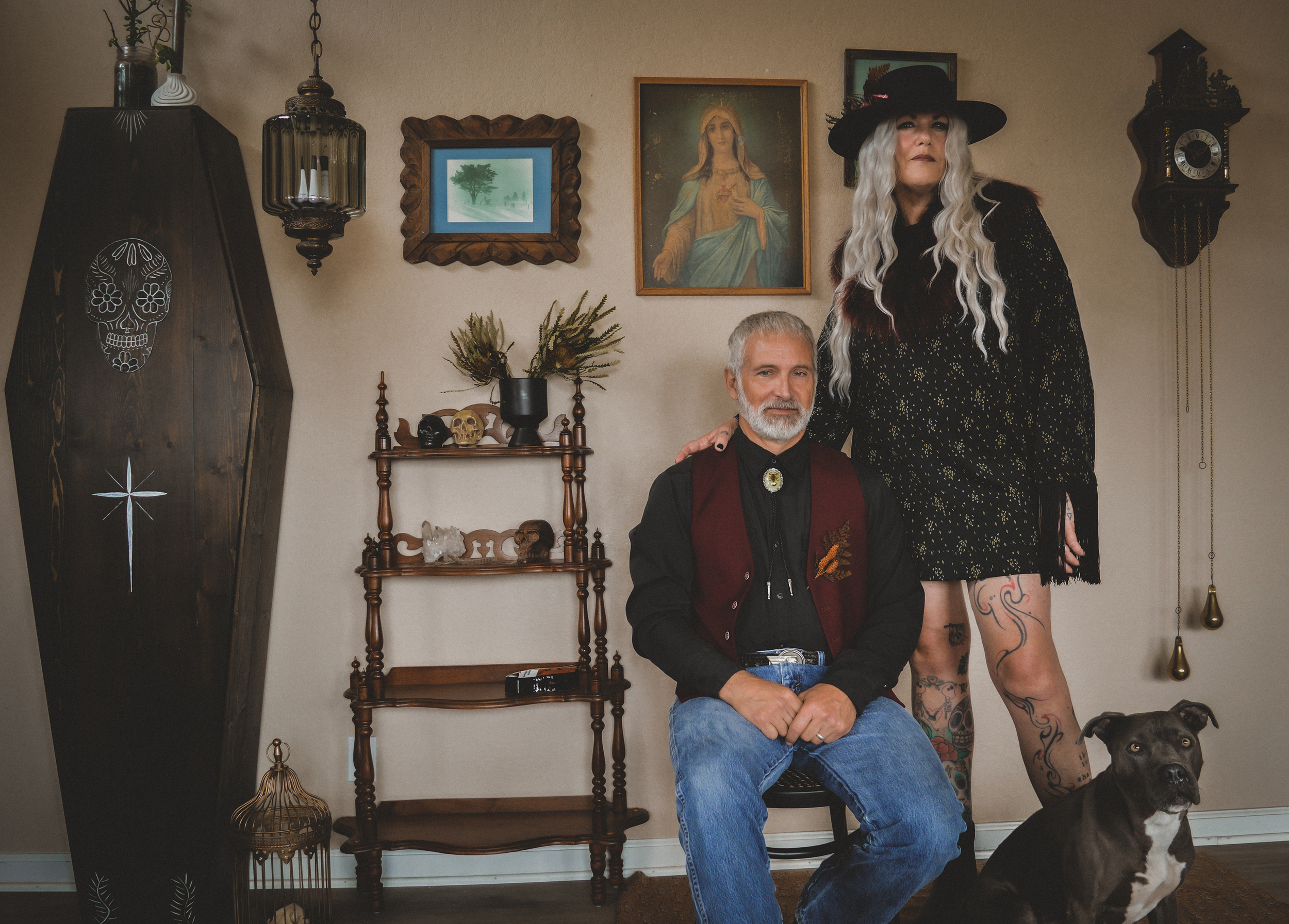 A woman with long silver hair and tattoos standing beside a seated man with a gray beard, in a room decorated with religious and vintage items. The woman is wearing a black hat and dress, and the man is wearing a black shirt, burgundy vest, and jeans