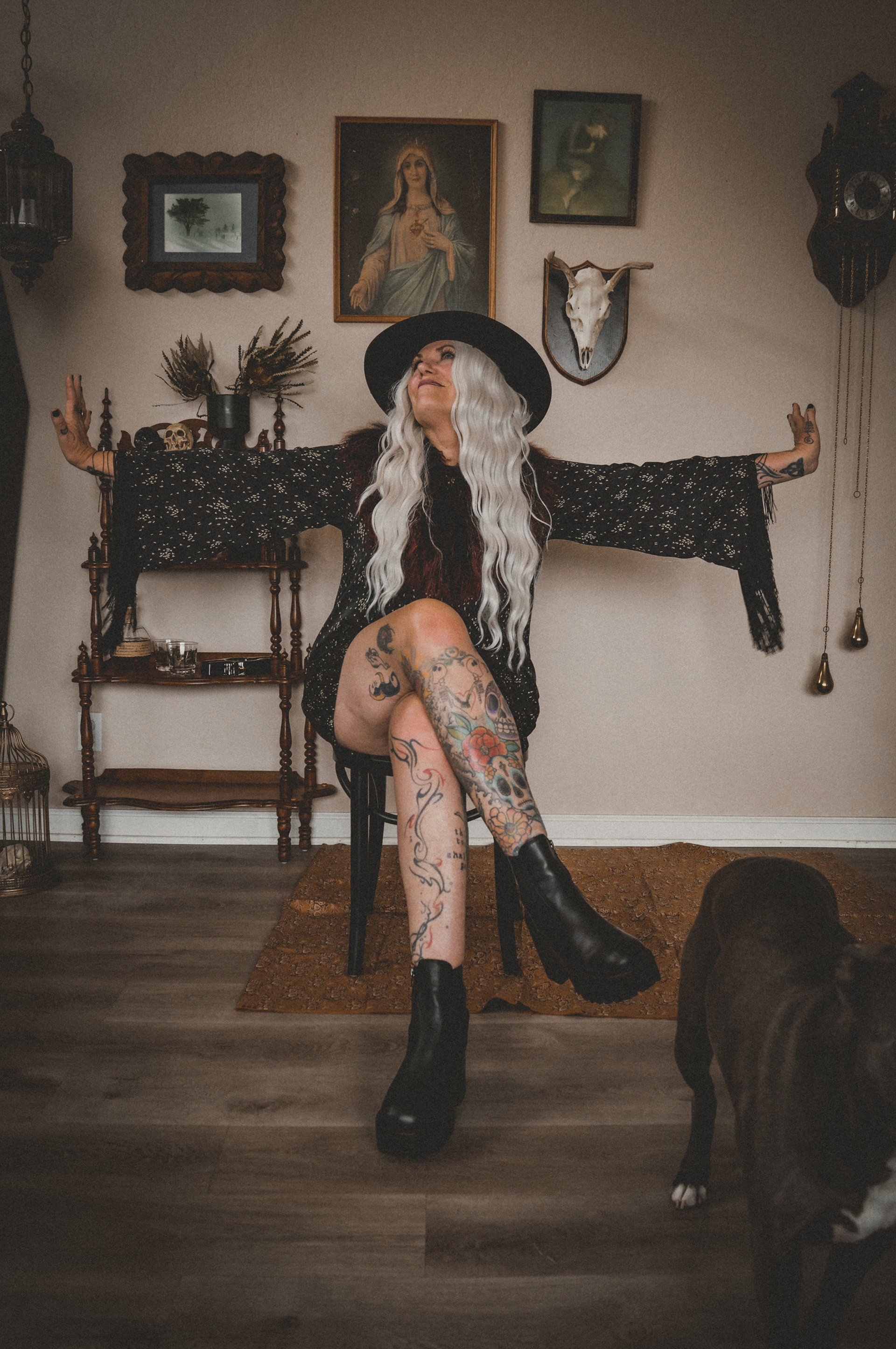 A woman with long, wavy silver hair wearing a black wide-brimmed hat, a black lace dress, and black platform boots, sitting on a chair with arms outstretched and smiling in a vintage-style room with wall art, a skull, and a dog.