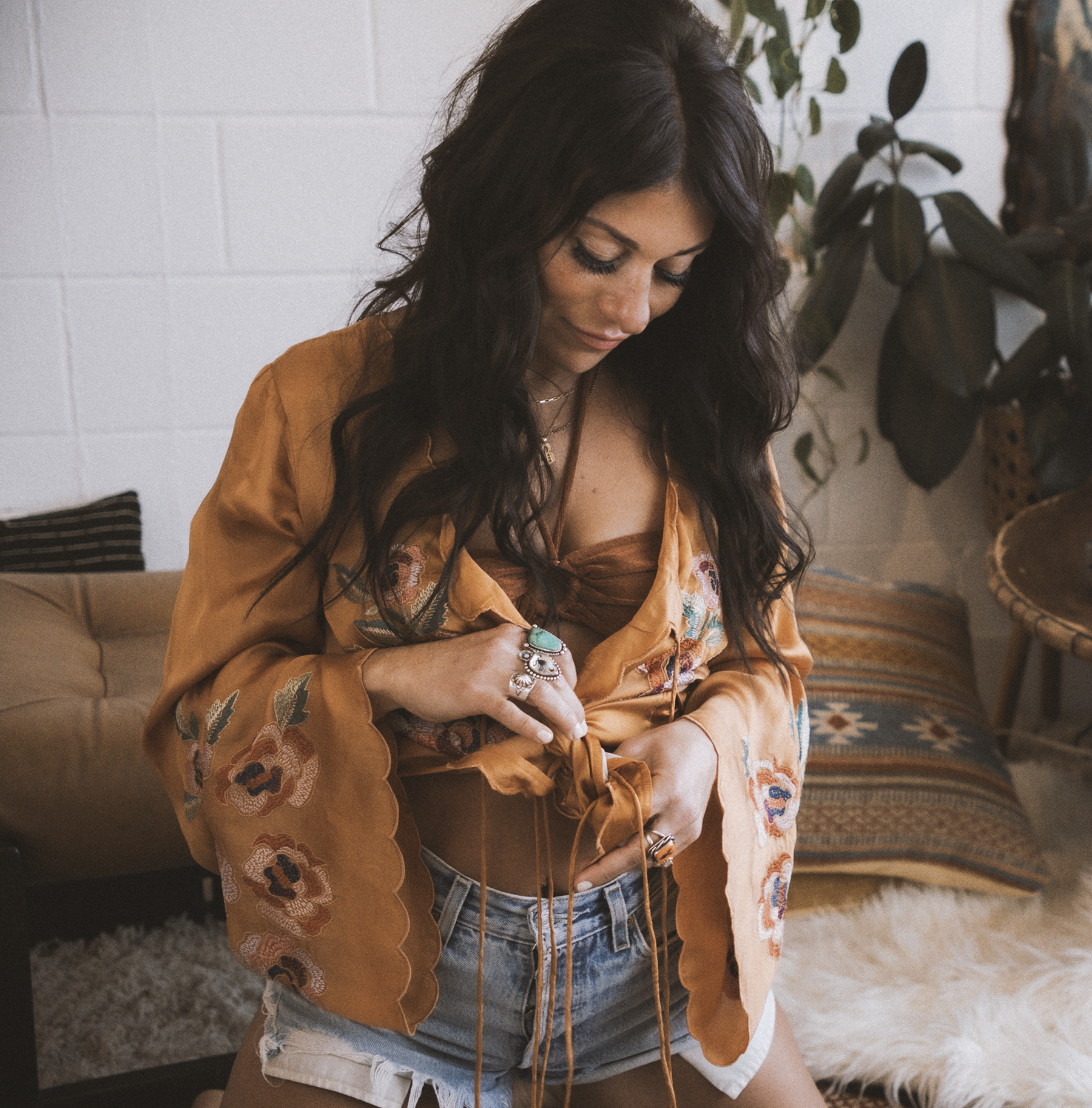A woman with long dark wavy hair wearing a satin embroidered brown and orange kimono-style robe, shorts, and multiple rings, is looking down as she adjusts her top in a cozy indoor setting with a beige couch, patterned pillows, a shaggy rug, and pott