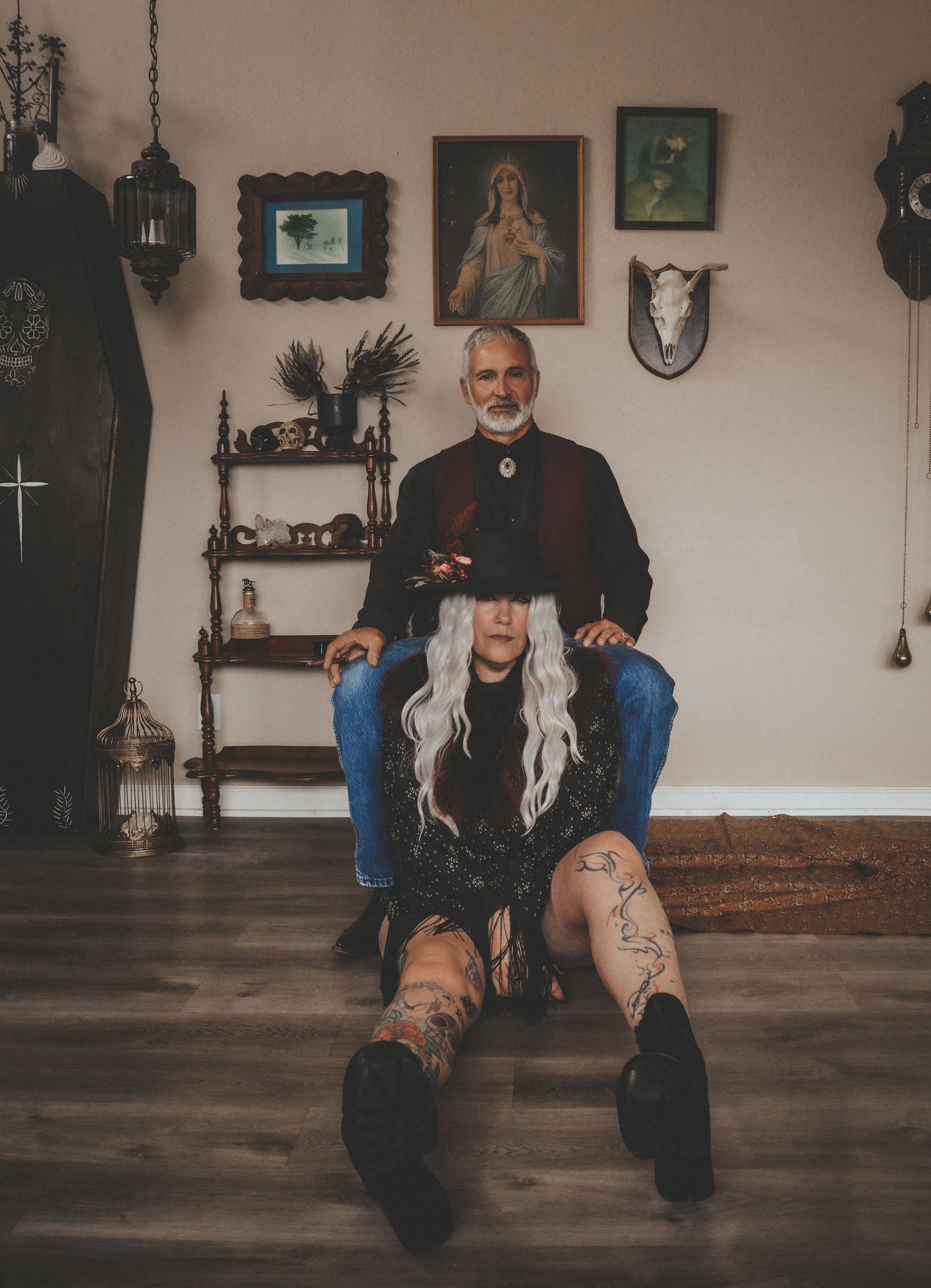 A man standing behind a woman sitting on the floor in a room with dark wood furniture and wall decorations, including a painting of a woman, a mounted animal skull, and framed artwork. The woman has long, wavy, platinum blonde hair, tattoos on her le