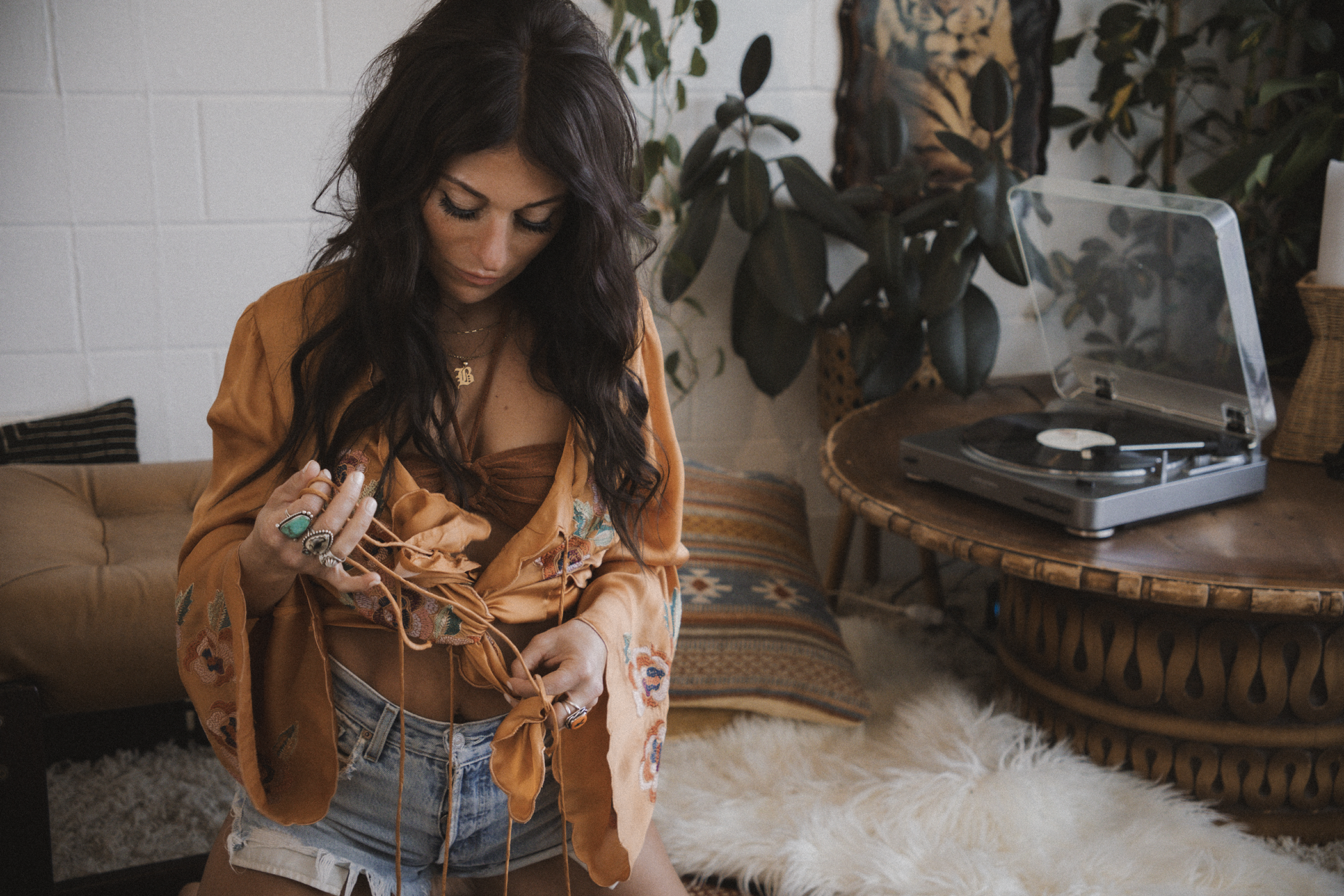 A young woman with long dark hair wearing a burnt orange floral silk robe and denim shorts is looking down and holding jewelry in her hands. She is in a room with a white brick wall, a rustic wooden table with a vintage turntable, a large leafy plant