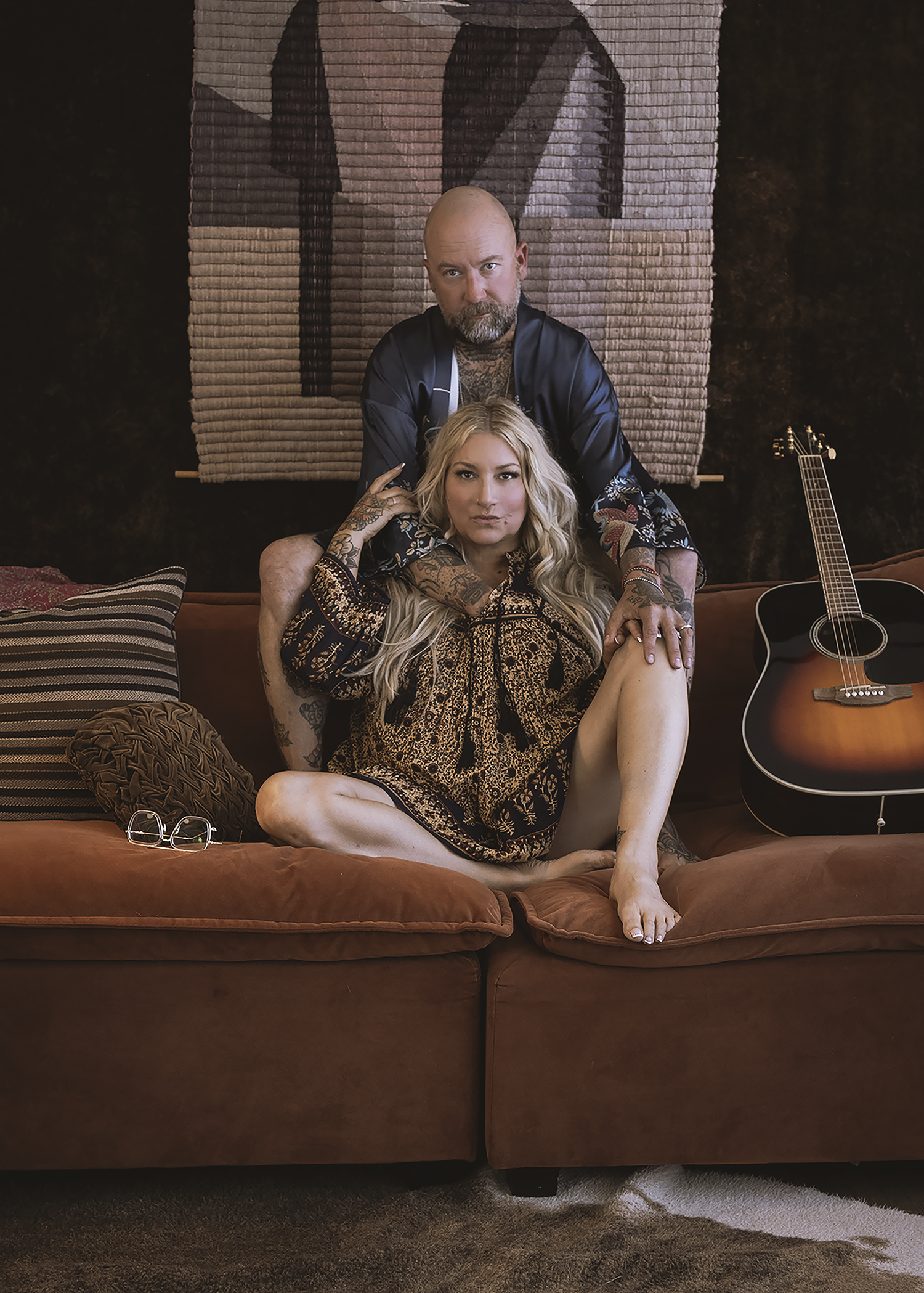 A man and woman with tattoos sit on a brown couch in a living room. The woman sits on the couch with her legs crossed, while the man is behind her, leaning on her shoulders. There are pillows on the couch and a guitar on the right side. A woven wall 