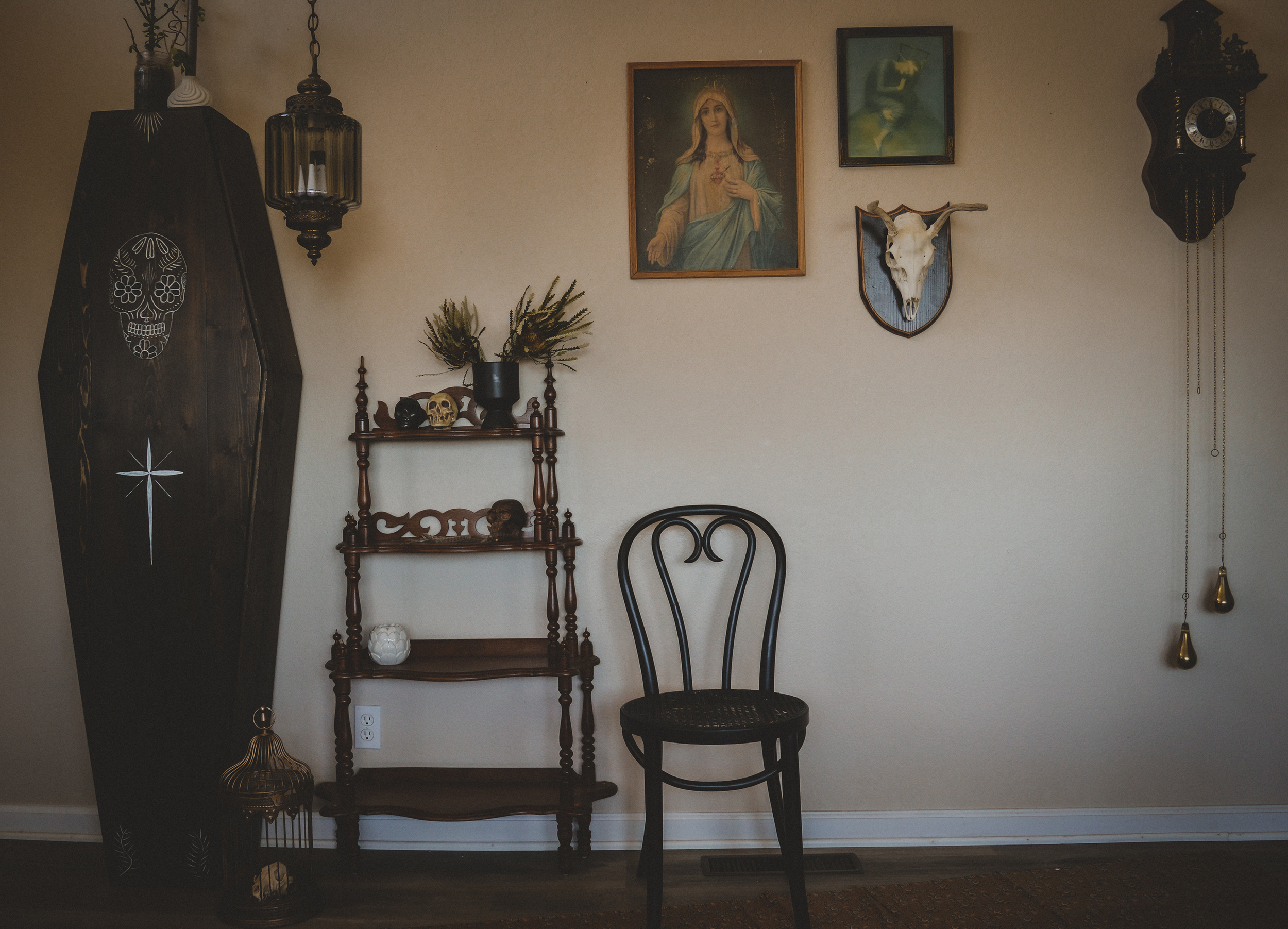Interior wall with decorative items, religious art, and vintage furniture including a black chair, wooden shelf with skull decorations, and a black coffin-style display with painted designs. Wall has religious and animal skull art, a clock, and hangi