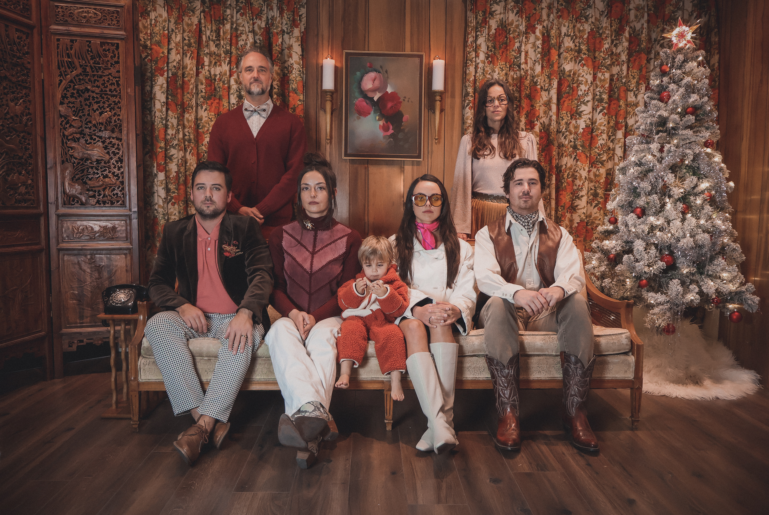 A group of nine people, including children, posing on a vintage sofa in a wood-paneled room decorated for Christmas with a large snow-frosted Christmas tree, floral painting, candles, and curtains.