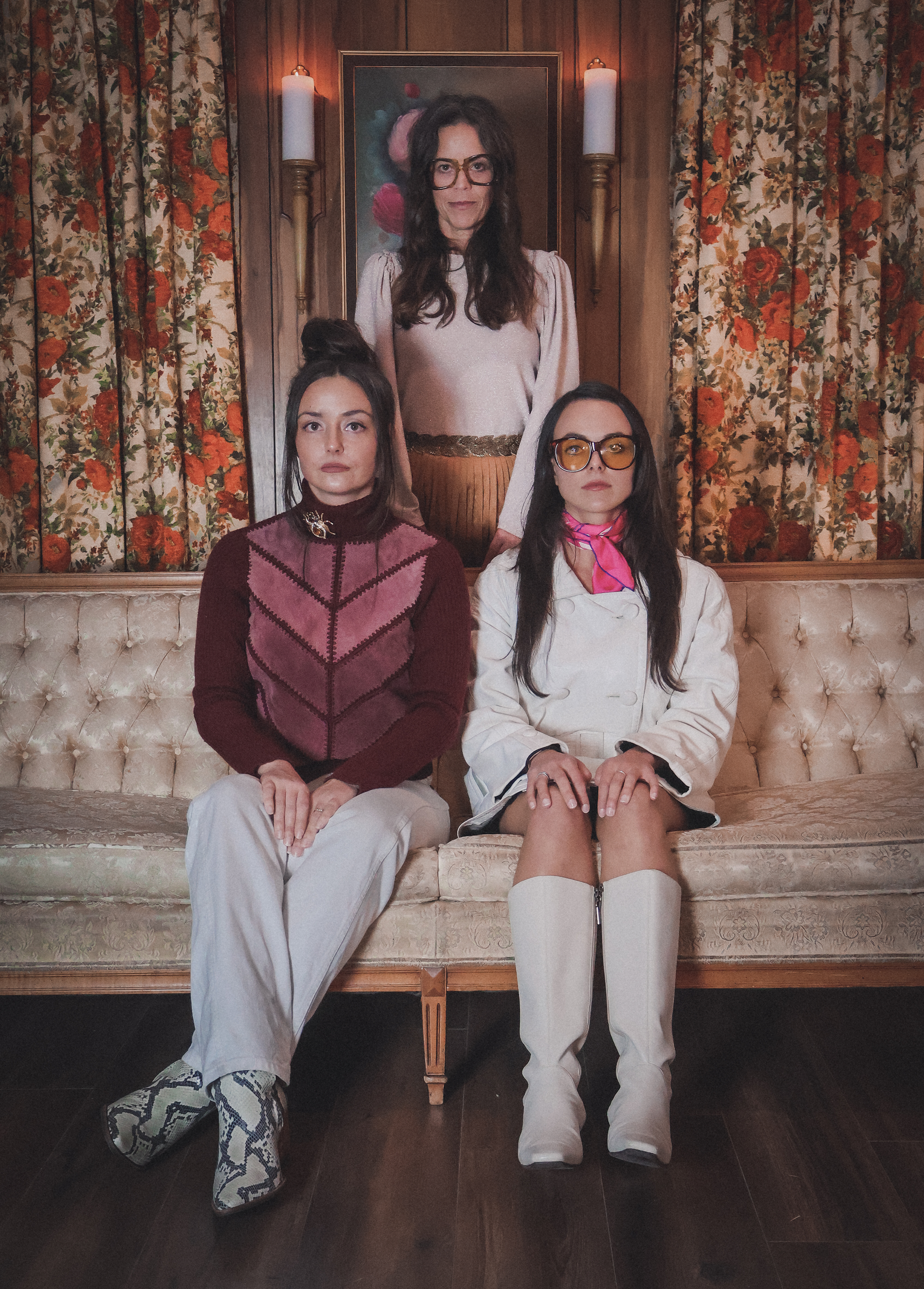 Three women sitting and standing in a vintage-style room with floral curtains. The woman at the back is standing, wearing glasses and a light-colored blouse. The woman on the left has her hair in a bun, wearing a burgundy and pink patterned sweater a