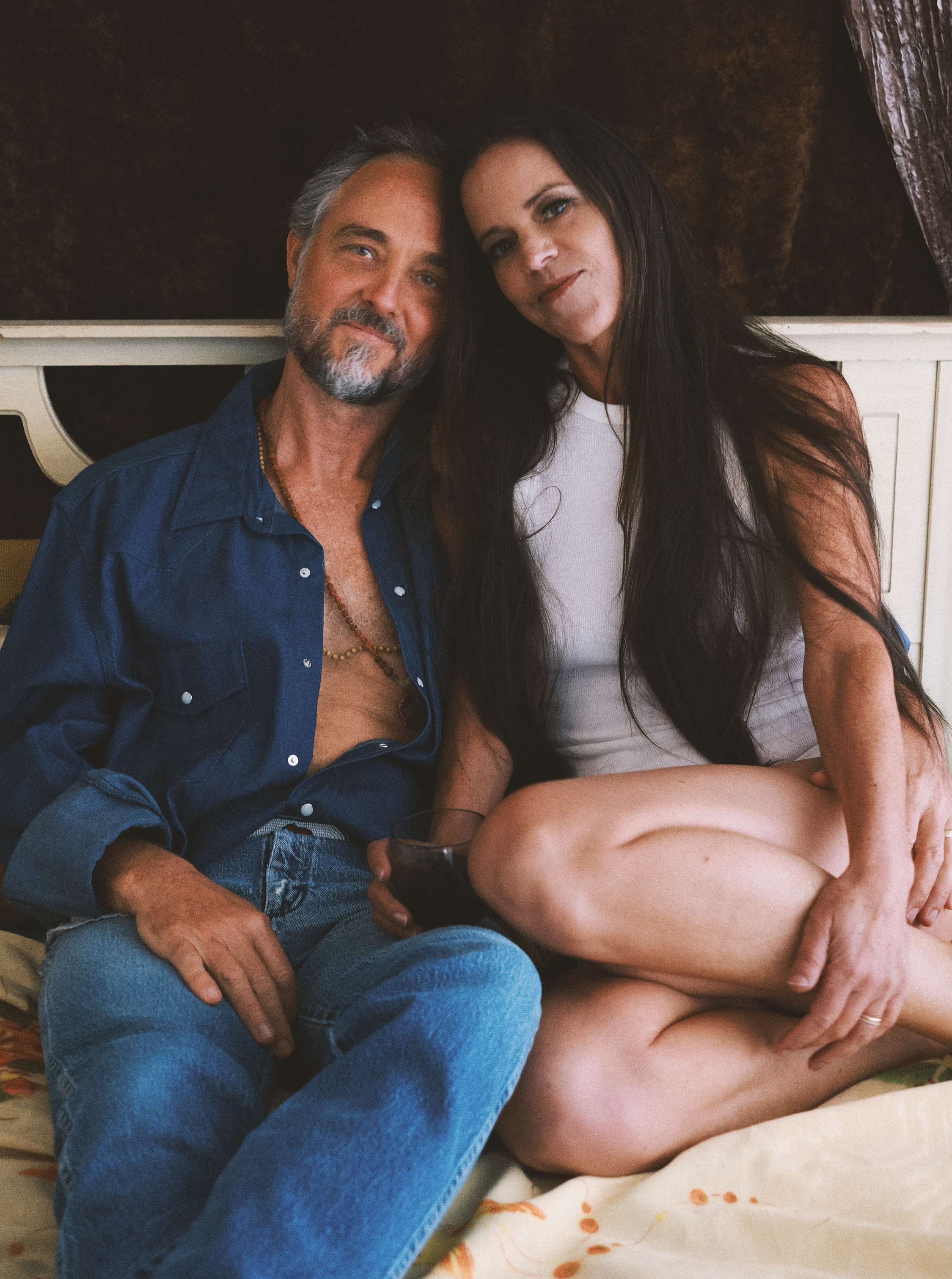 A man and a woman sitting together on a bed, smiling at the camera. The man has gray hair, a beard, and is wearing a blue denim shirt. The woman has long dark hair, wearing a white top, and holding a glass of red wine. They are seated closely with th
