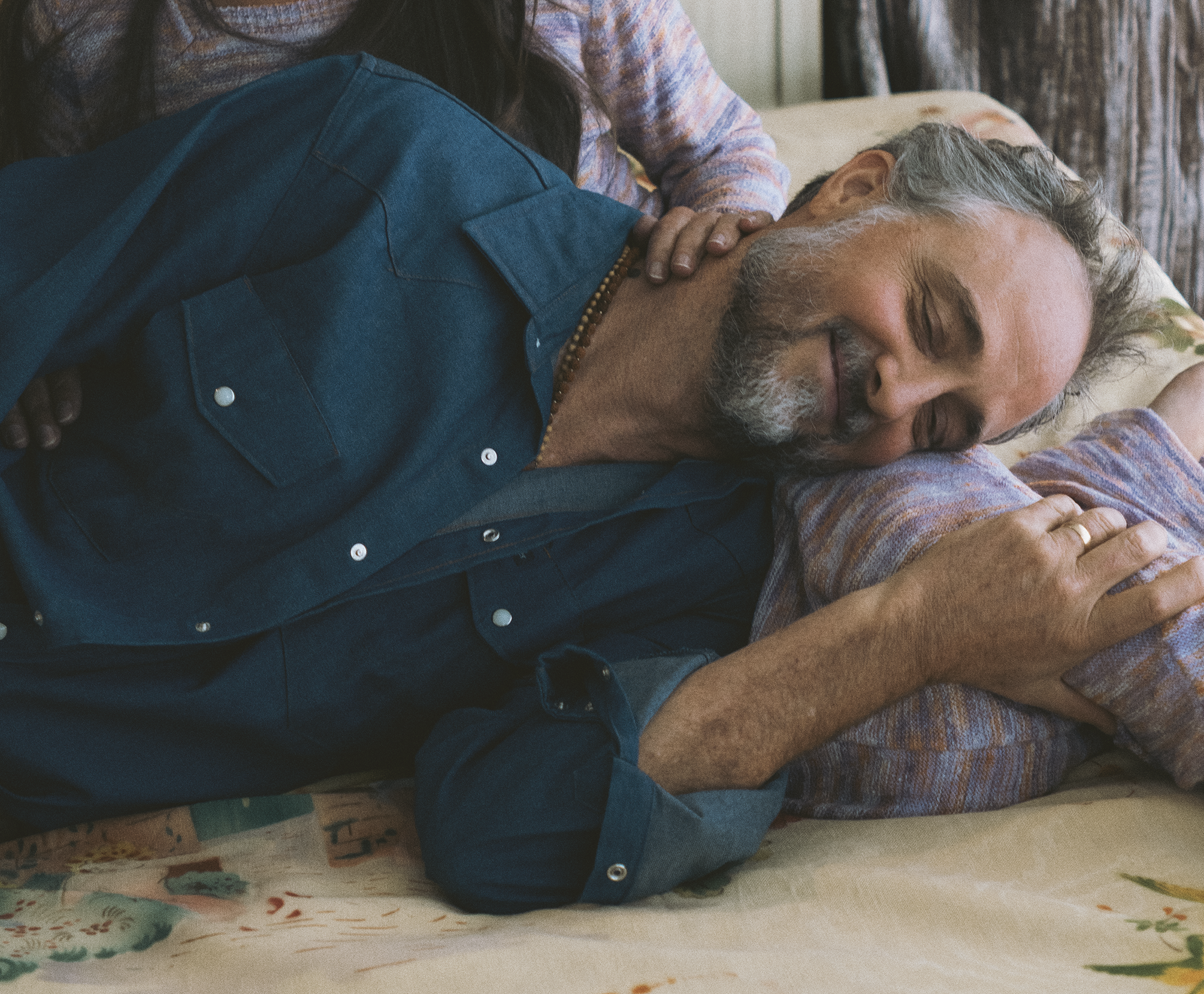 A senior man with gray hair and beard, smiling peacefully while resting on a bed with his eyes closed. A woman with long dark hair and hands on his shoulder is leaning over him in a caring manner.