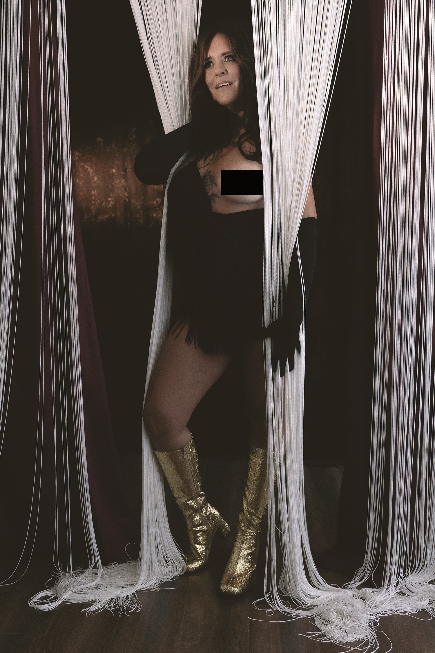 Woman dressed in black with gold boots standing among strings or threads hanging from above and around her.