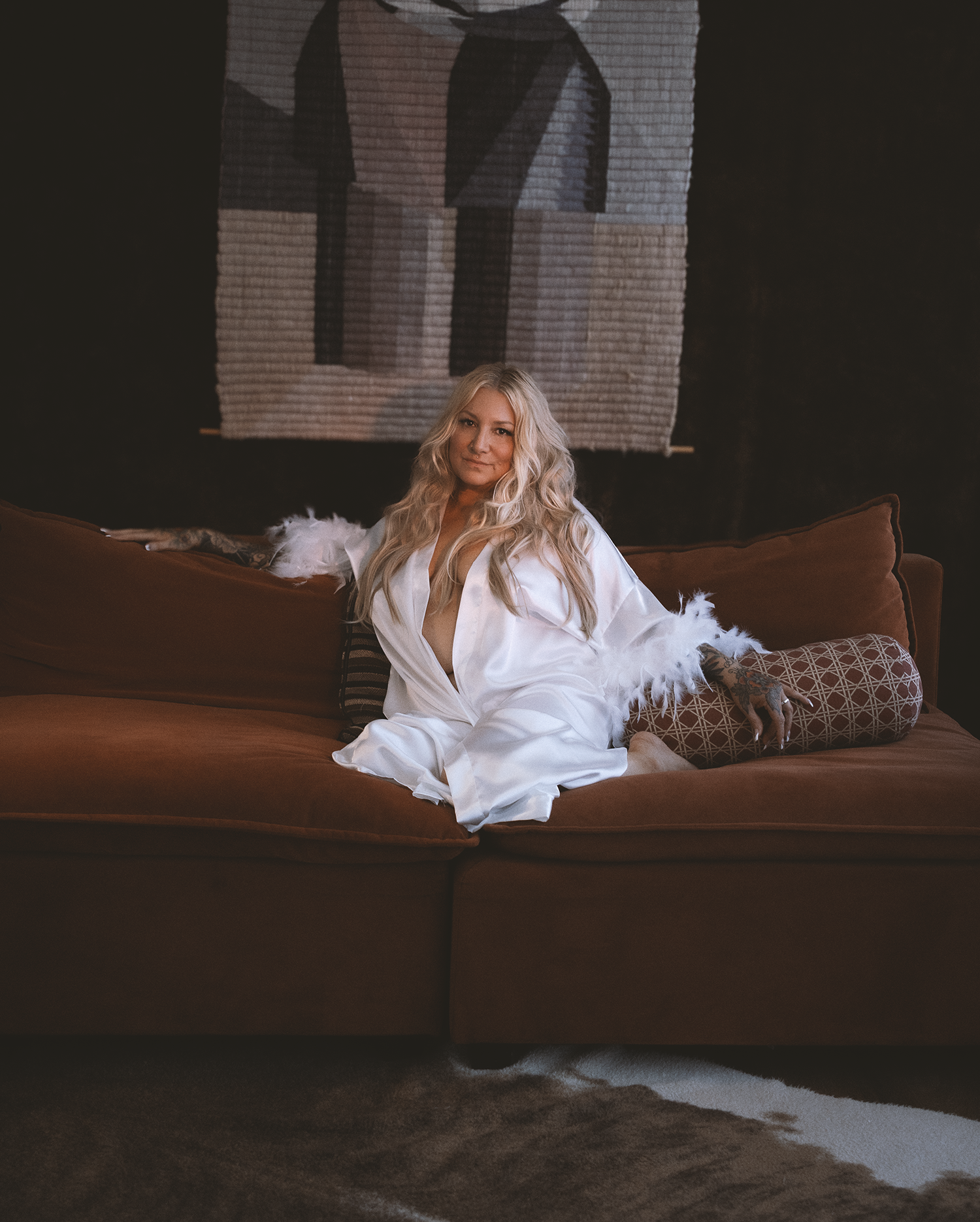 A woman with long blonde hair wearing a white satin robe with feathered sleeves sitting on a brown sofa in a dimly lit room with a decorative tapestry in the background.