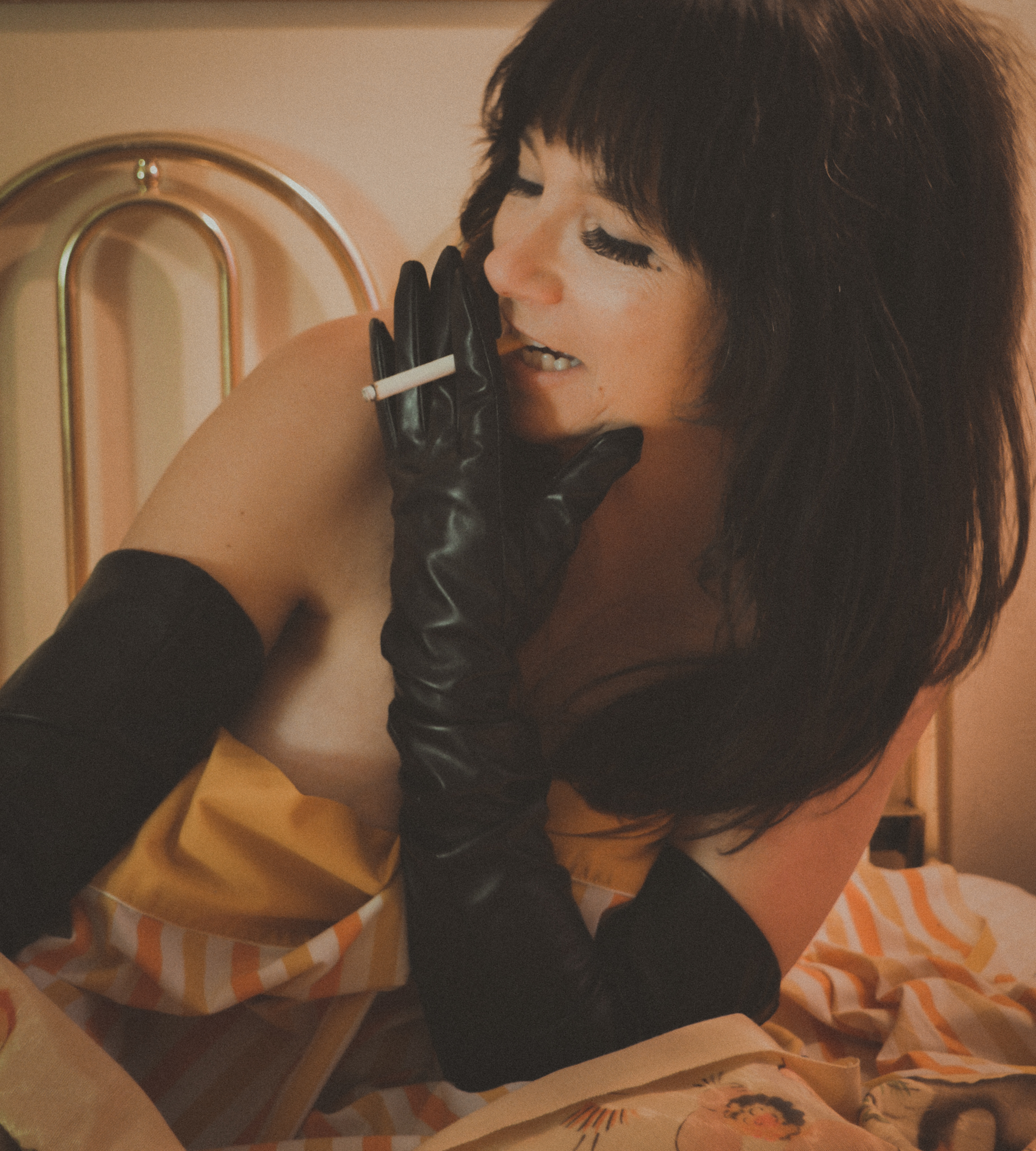 A woman with black hair and makeup, wearing black gloves and a striped yellow and beige dress, sitting on a bed with a striped bedspread, holding a cigarette in her gloved hand and leaning forward, with a headboard visible in the background.