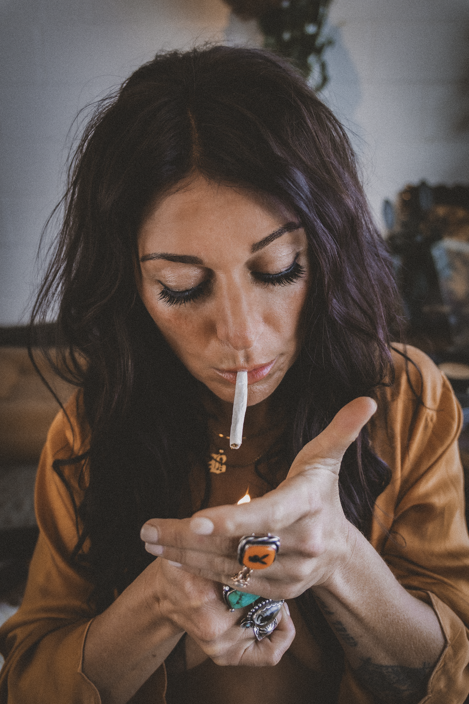 A woman with dark, wavy hair and long eyelashes is lighting a cigarette with a match, wearing rings and a necklace.