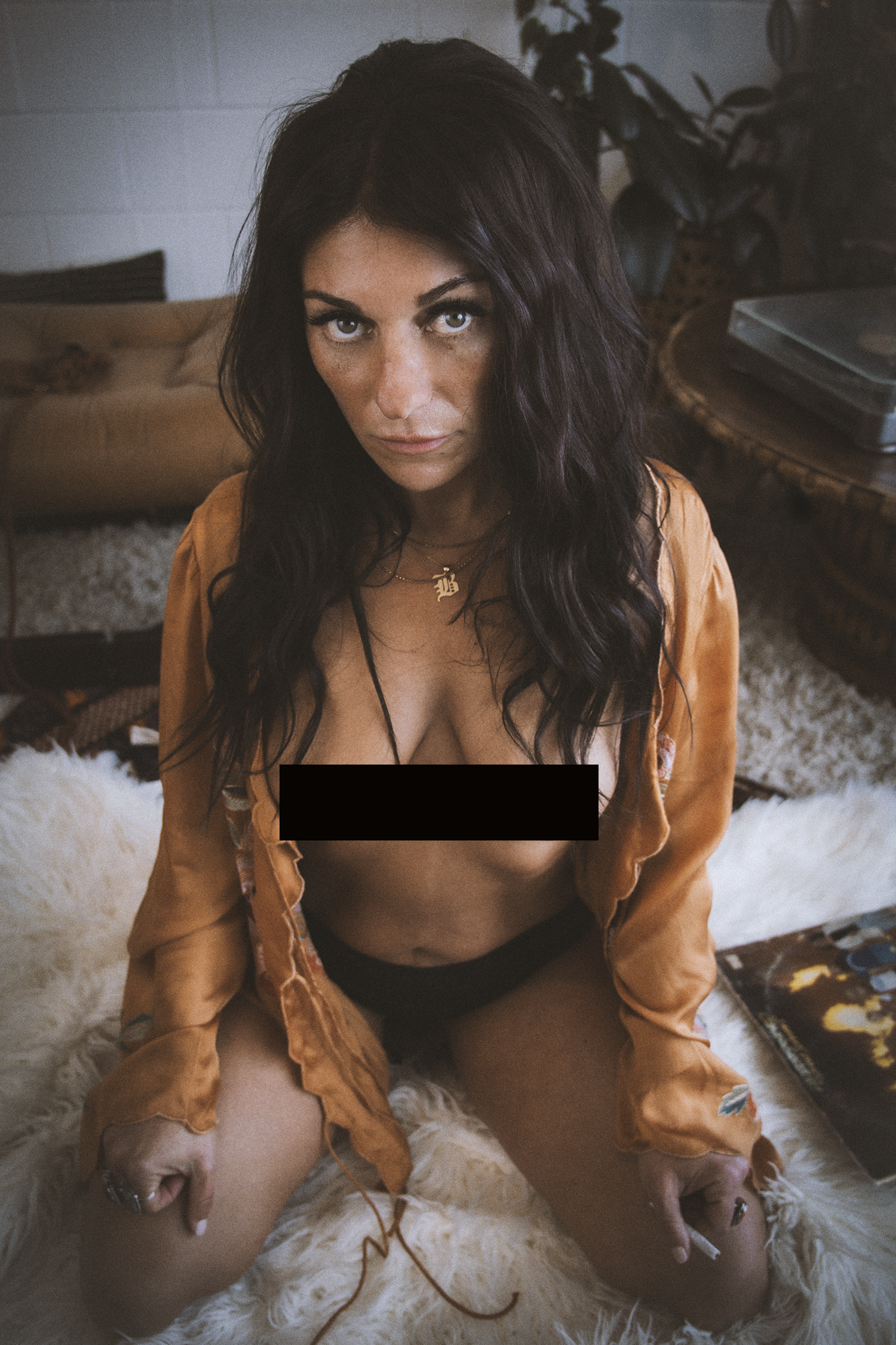 A woman with long dark wavy hair, wearing a gold satin robe and a necklace with a pendant, sits on a fluffy white rug in a room with a sofa, plants, and a shelf with books or albums. She looks directly into the camera with an intense expression.