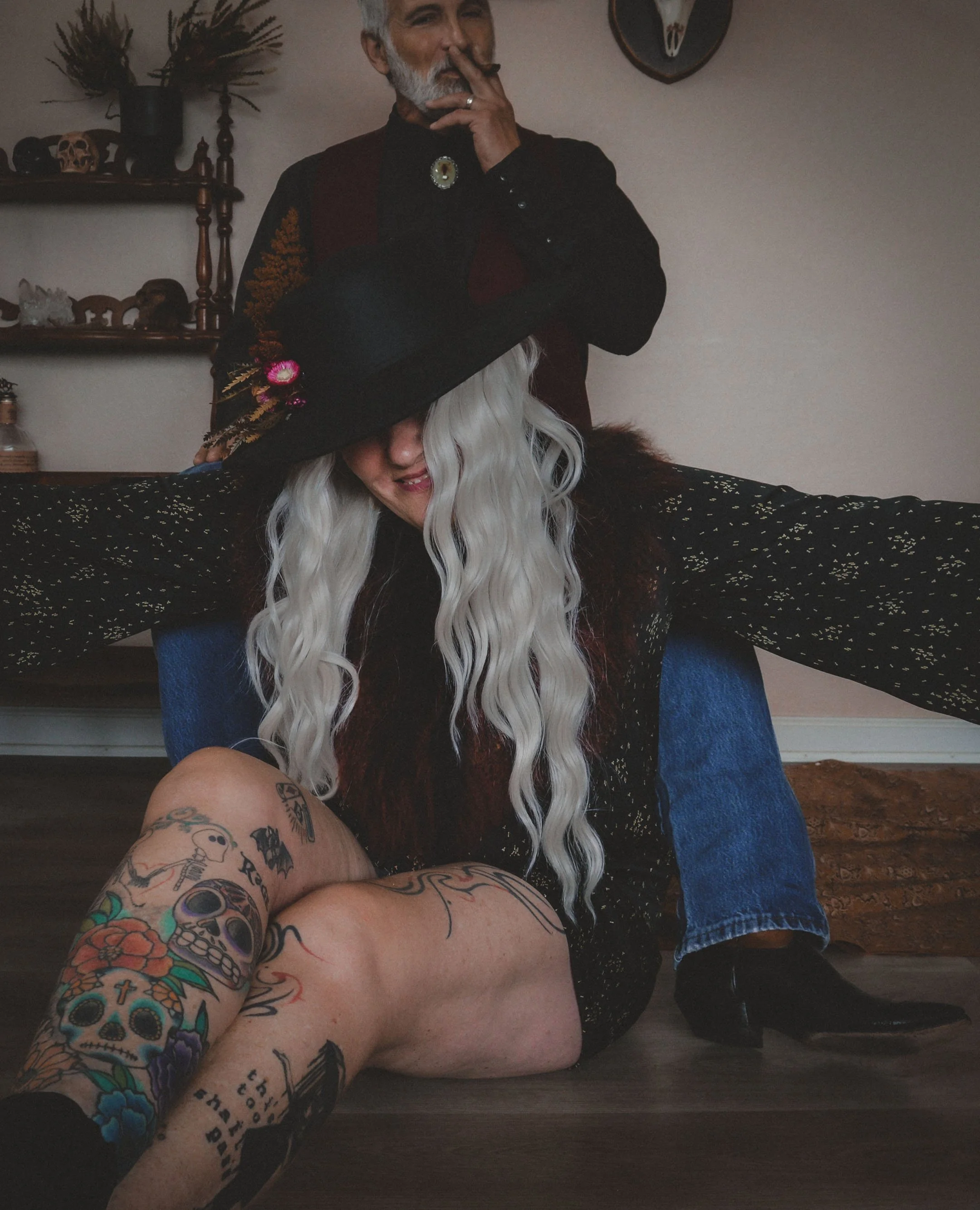 A woman with long, wavy platinum blonde hair wearing a large black hat with colorful decorations, sitting cross-legged on the floor with her arms outstretched, and a man standing behind her with his hand near his face. The woman has tattoos on her le