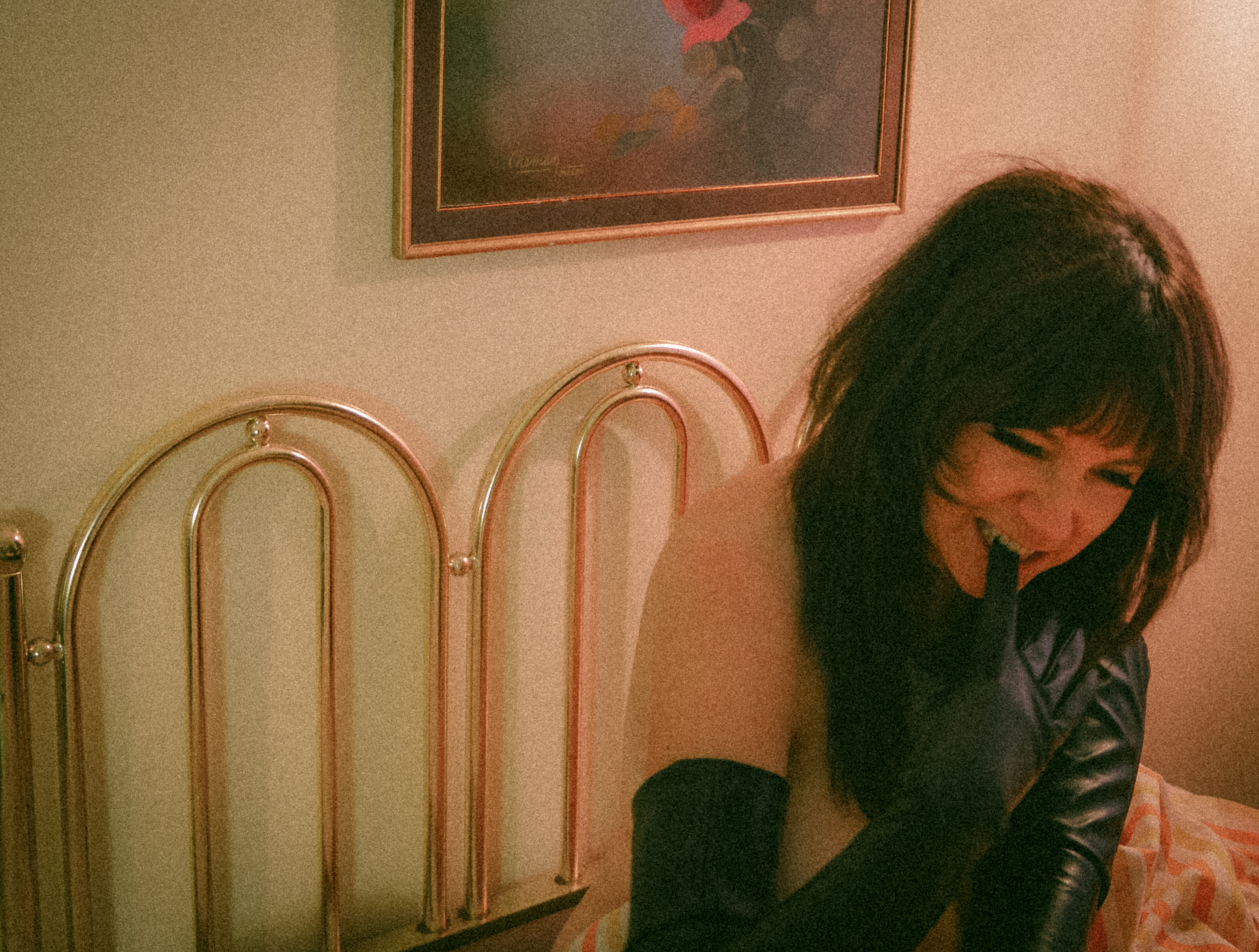 A woman with dark hair smiling, holding a black object in her mouth, wearing a sleeveless top and black gloves, sitting on a bed with a striped bedsheet in a room with a pink and green wall, a framed floral painting, and a golden metal headboard.