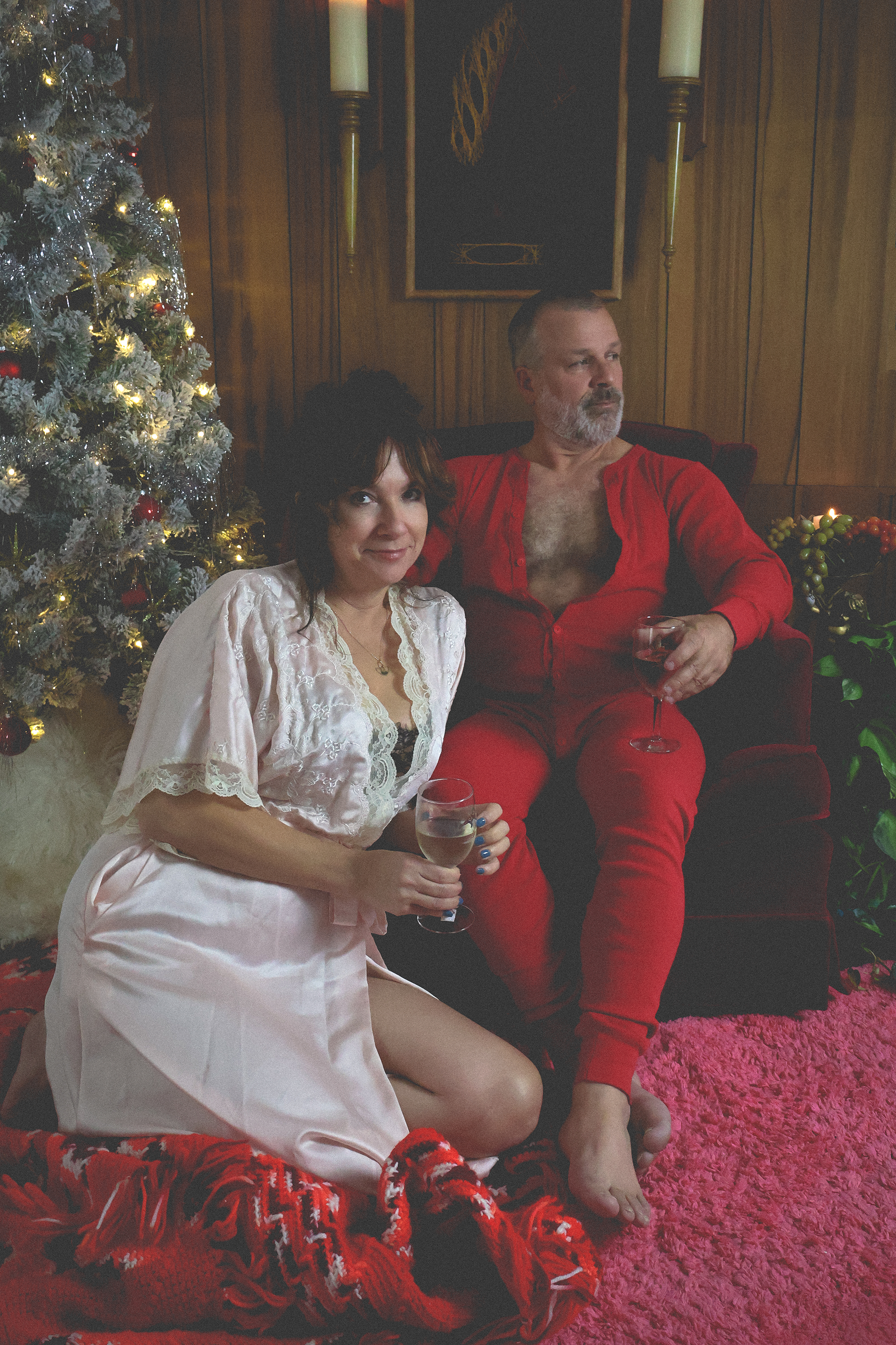 A woman and a man sitting on a pink carpet beside a decorated vintage Christmas tree, holding glasses of wine, in a cozy, wood-paneled room with Christmas decorations. 