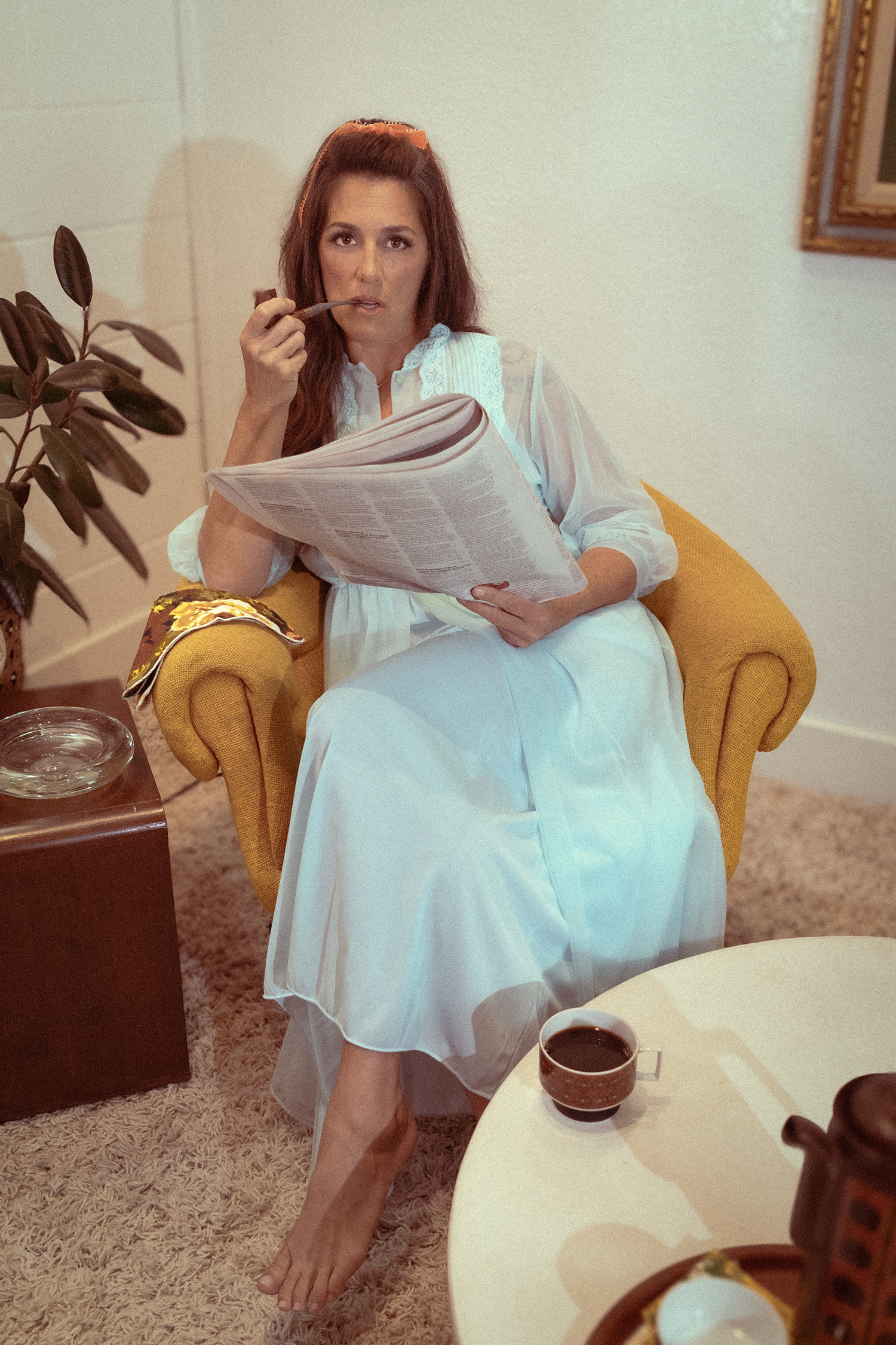 A woman with brown hair sitting in a yellow armchair, wearing a white dress, holding a newspaper, and drinking coffee from a patterned mug on a white table. She is looking at the camera with a thoughtful expression.