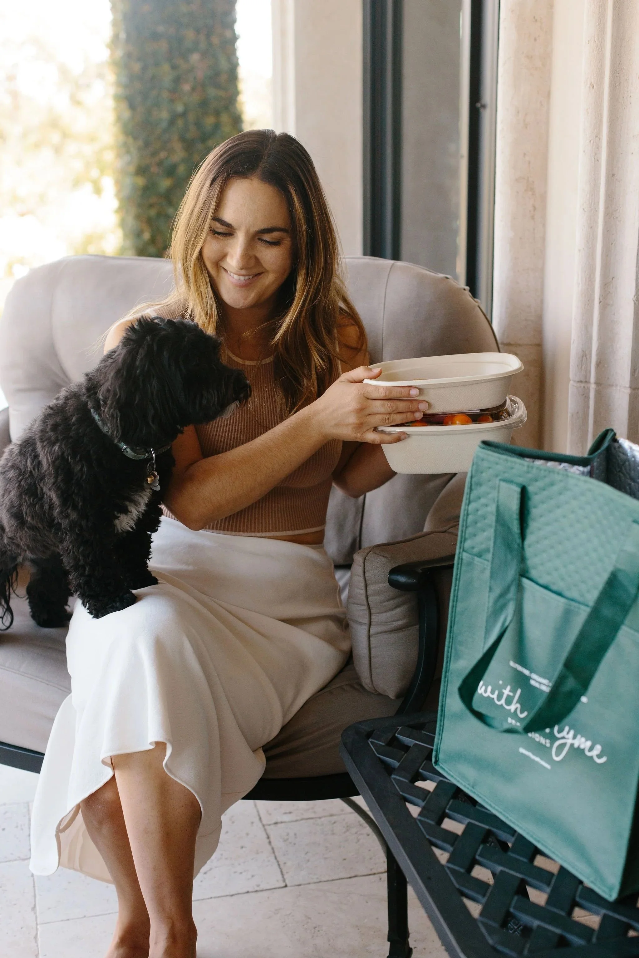 A woman sitting on a cushioned chair, holding With Thyme Provisions food containers, with a small black dog on her lap, smiling indoors near a large window.