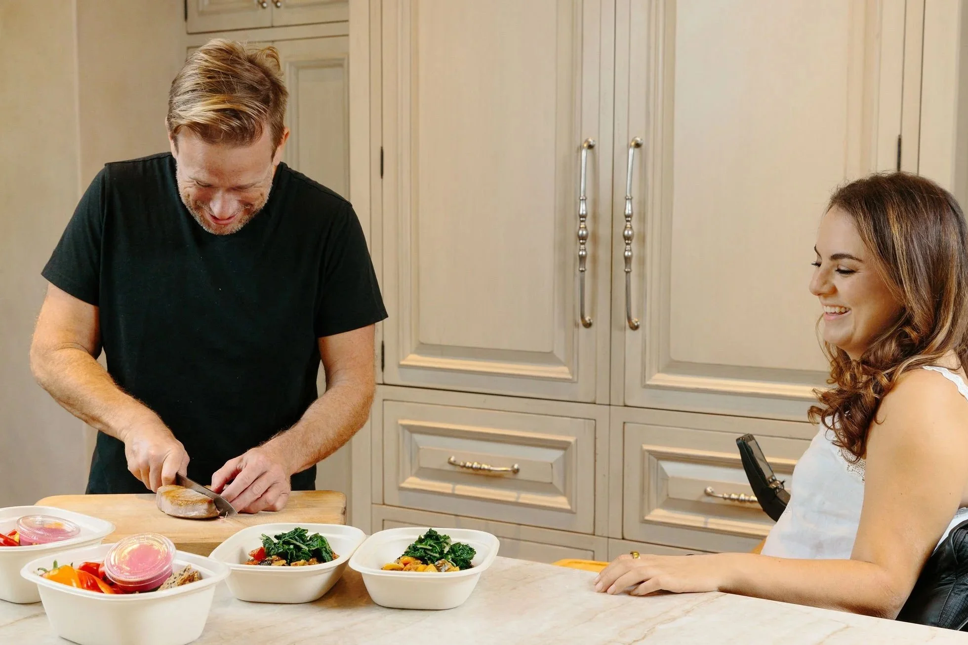 Chef is chopping food in a kitchen, smiling and looking down. Alanna is sitting at the kitchen counter, smiling and looking at the man. There are bowls of salad and other food items on the counter.