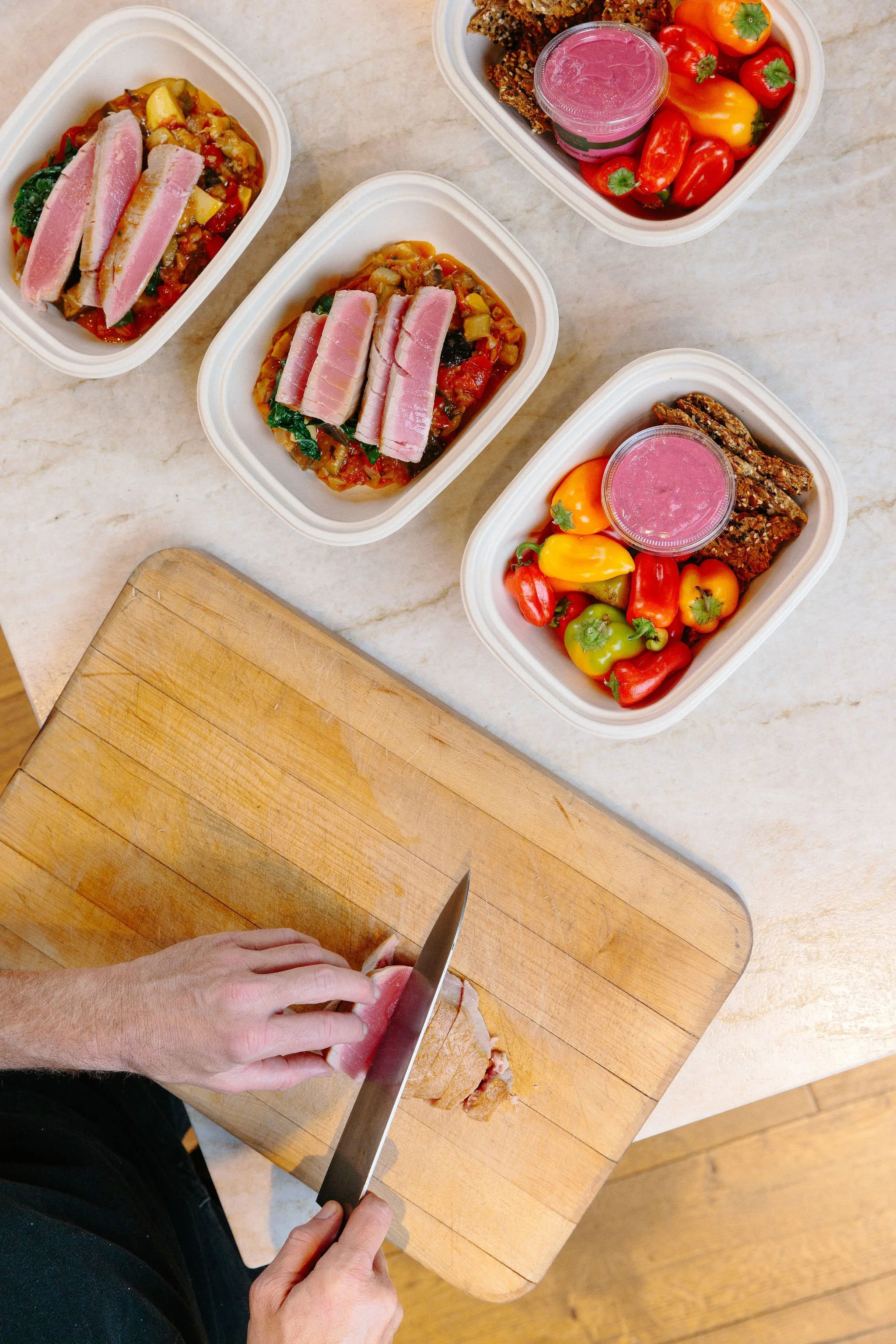 Person slicing cooked meat on a wooden cutting board, with three meal containers and assorted colorful peppers and dips on a light countertop.