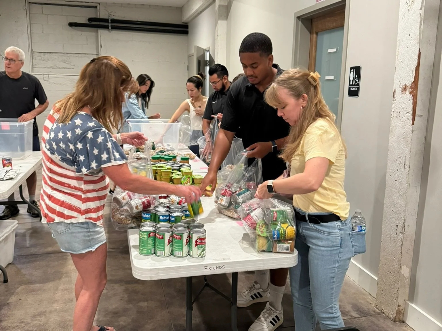 People packing food items into bags at a long table in a community center or similar environment.