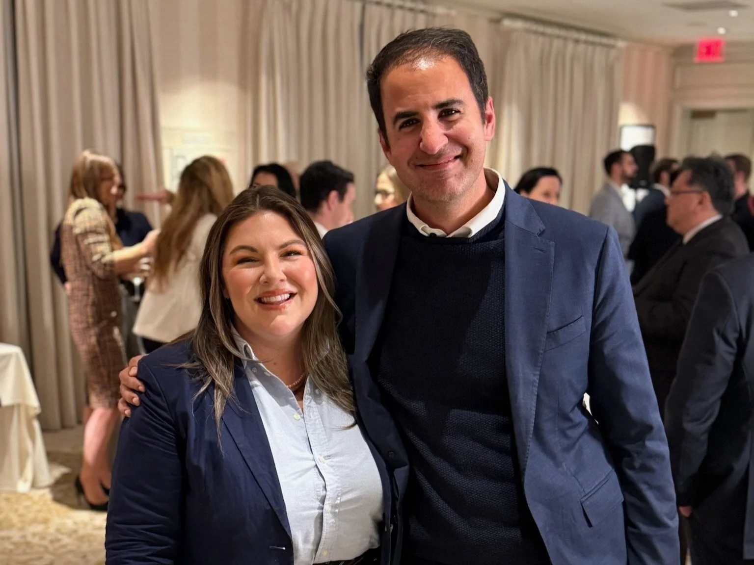 A woman and a man smiling and posing for a photo at an indoor event, with several people talking in the background.
