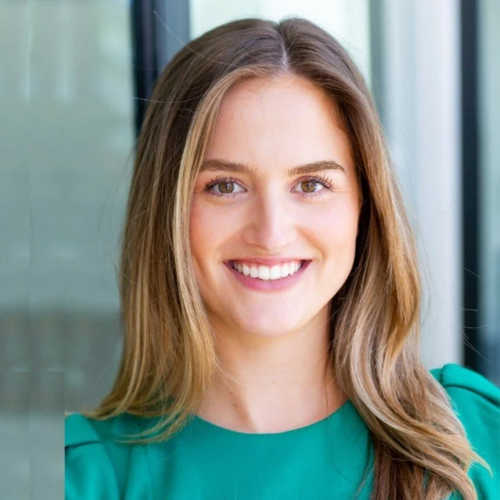 A young woman with long brown hair and light skin smiling, wearing a teal shirt, standing indoors near a glass window.