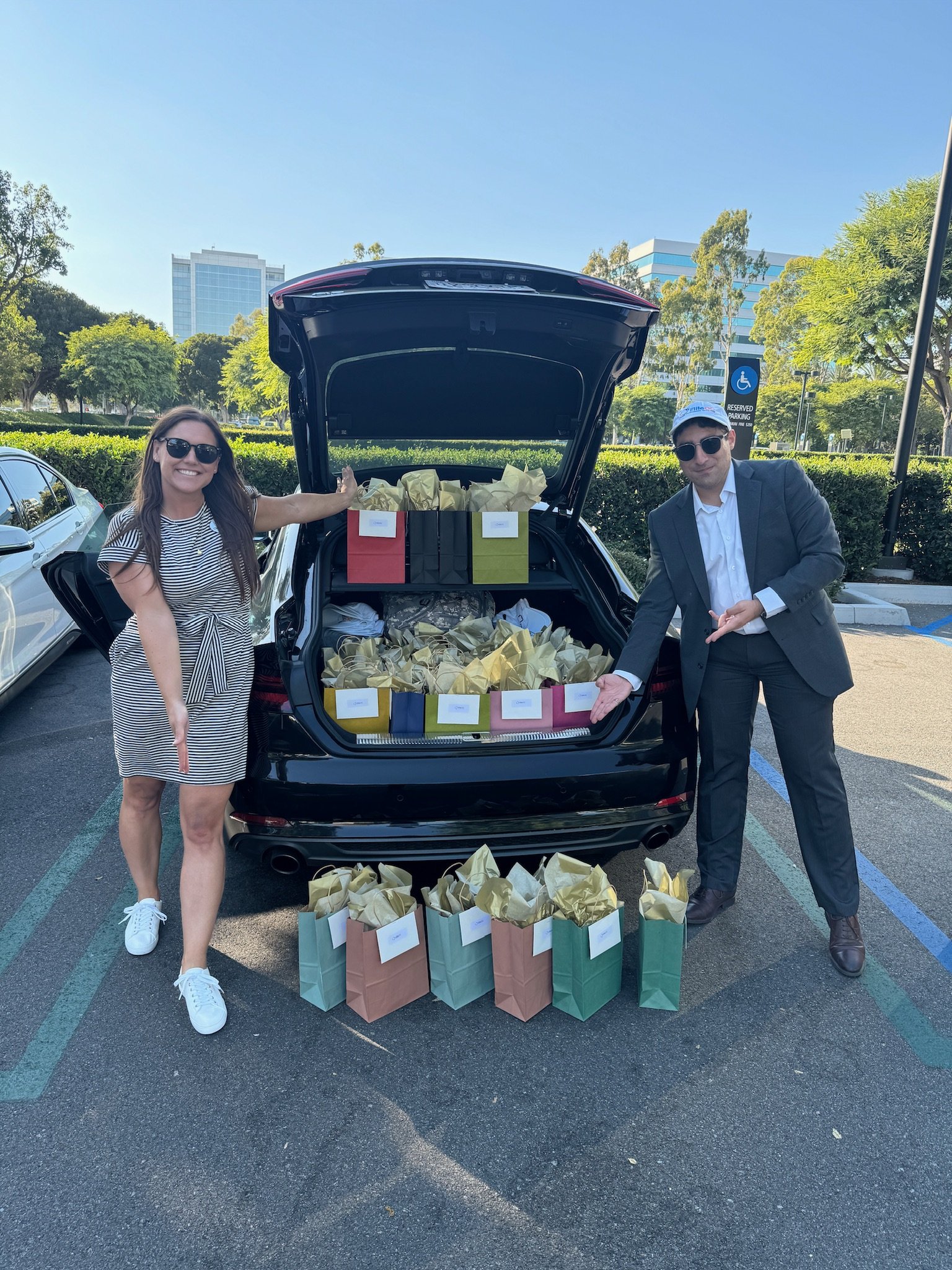 A woman and a man standing beside an open car trunk filled with gift bags in a parking lot, with a reserved space and city buildings in the background.