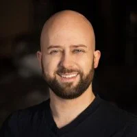 A man with a bald head and a beard smiling, wearing a black shirt.