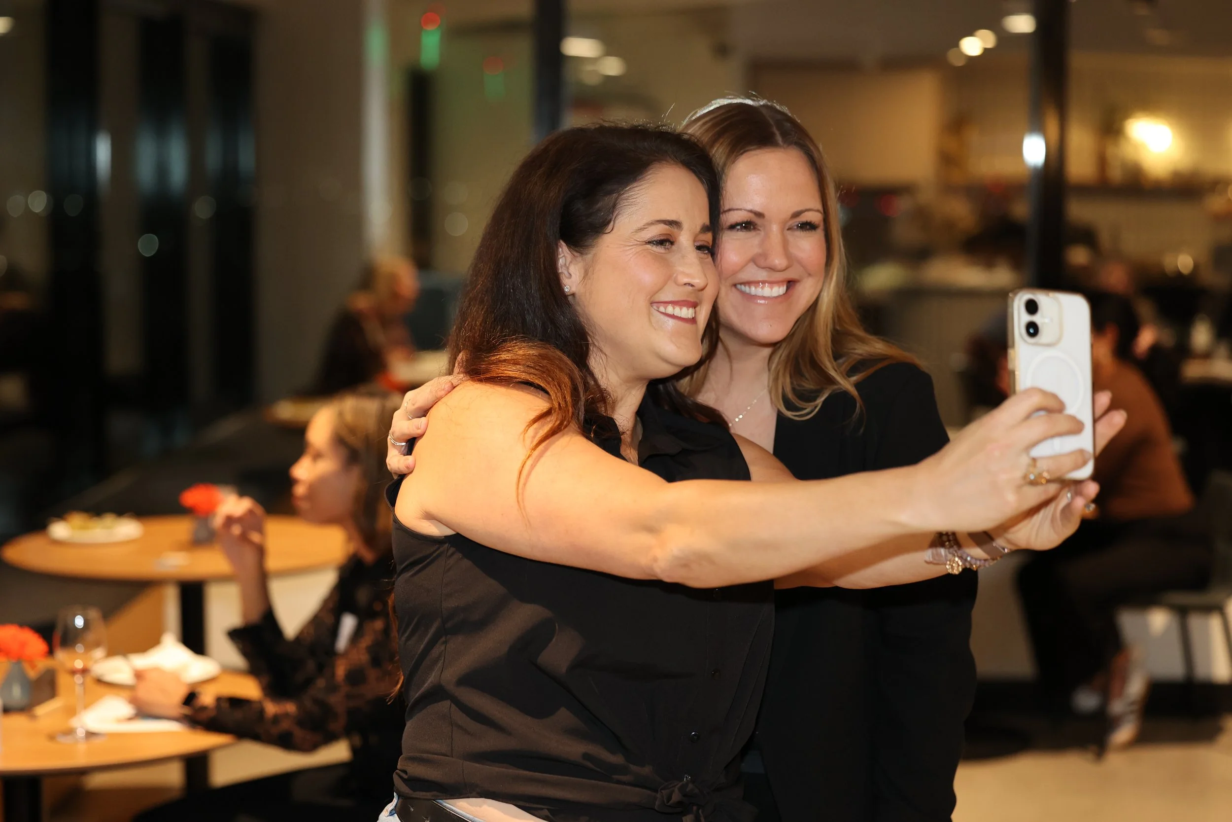 Two women are smiling and taking a selfie together in a restaurant or café setting, with one woman in a black top and the other in a dark dress.