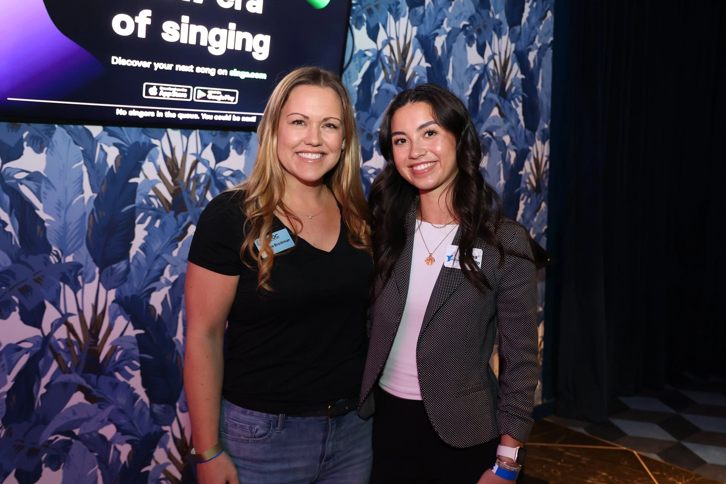 Two women standing side by side, smiling at the camera in front of a blue and white leafy patterned wall, with a large screen displaying 'Discover your next song' in the background.
