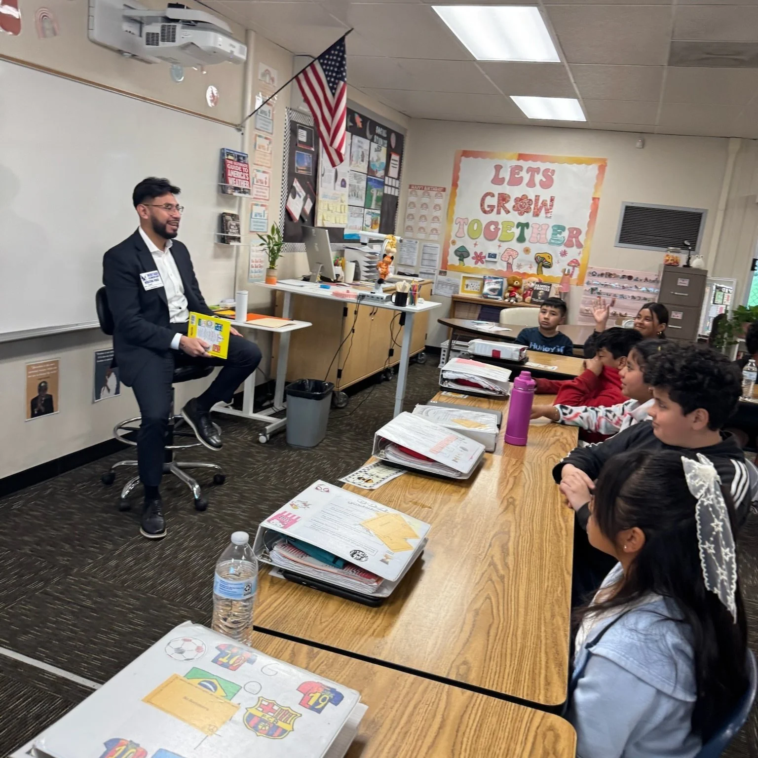 A male teacher sitting on a stool and holding a book in a classroom, with students sitting at their desks raising their hands. The classroom has educational posters, a whiteboard, and a large colorful sign that reads 'Let's Grow Together' on the wall.