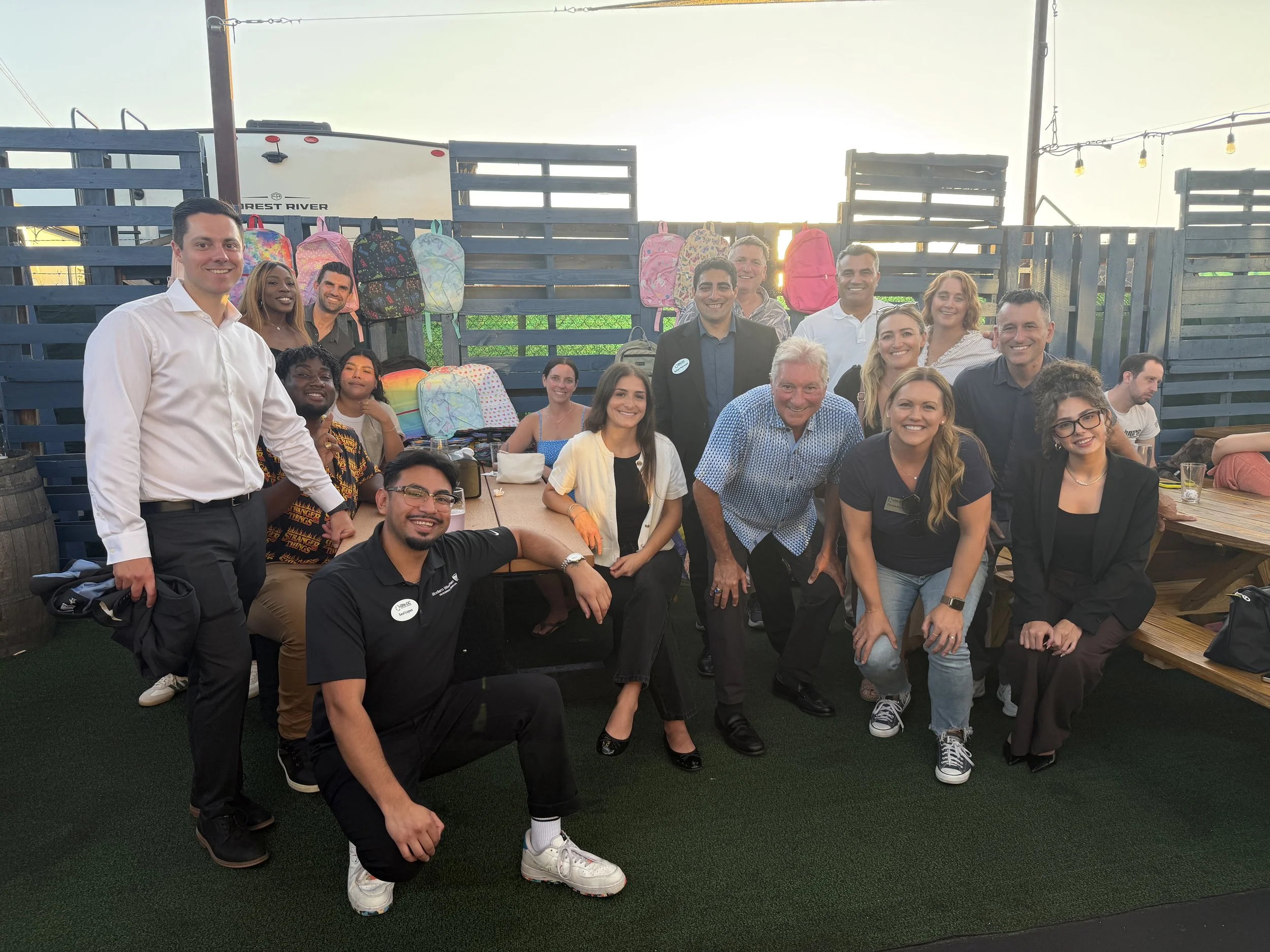 Group of people smiling and gathered at an outdoor event, with backpacks hanging on a wooden fence behind them. The scene is during sunset with string lights overhead.