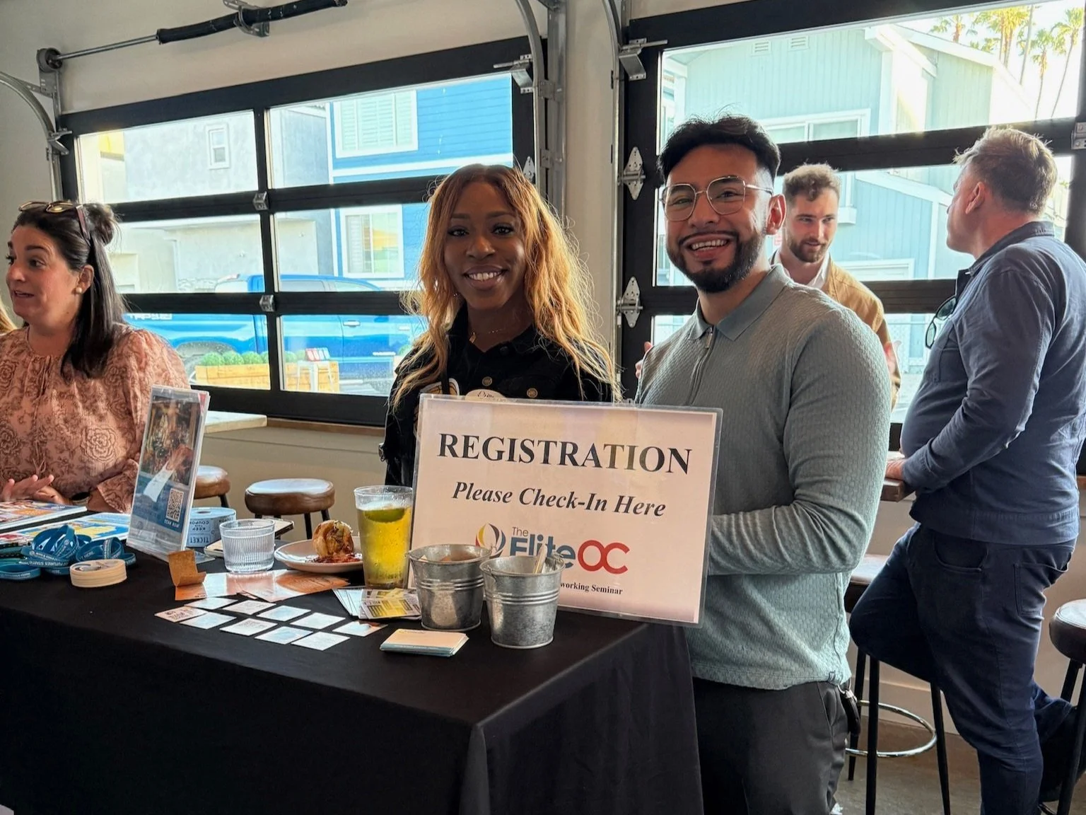 Two people smiling and holding a sign that reads 'Registration, Please Check-In Here' at an event registration table by a window, with other attendees in the background.