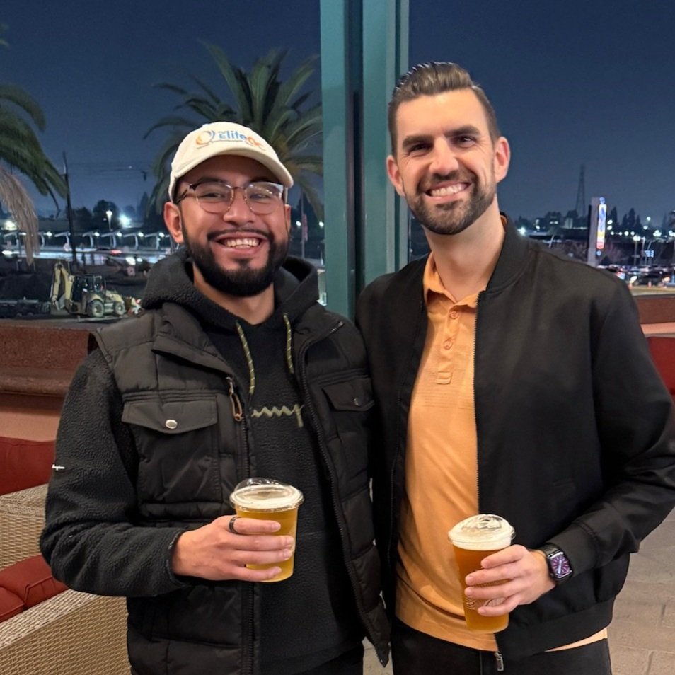 Two smiling men holding cups of beer, standing together outside at night with palm trees and city lights in the background.