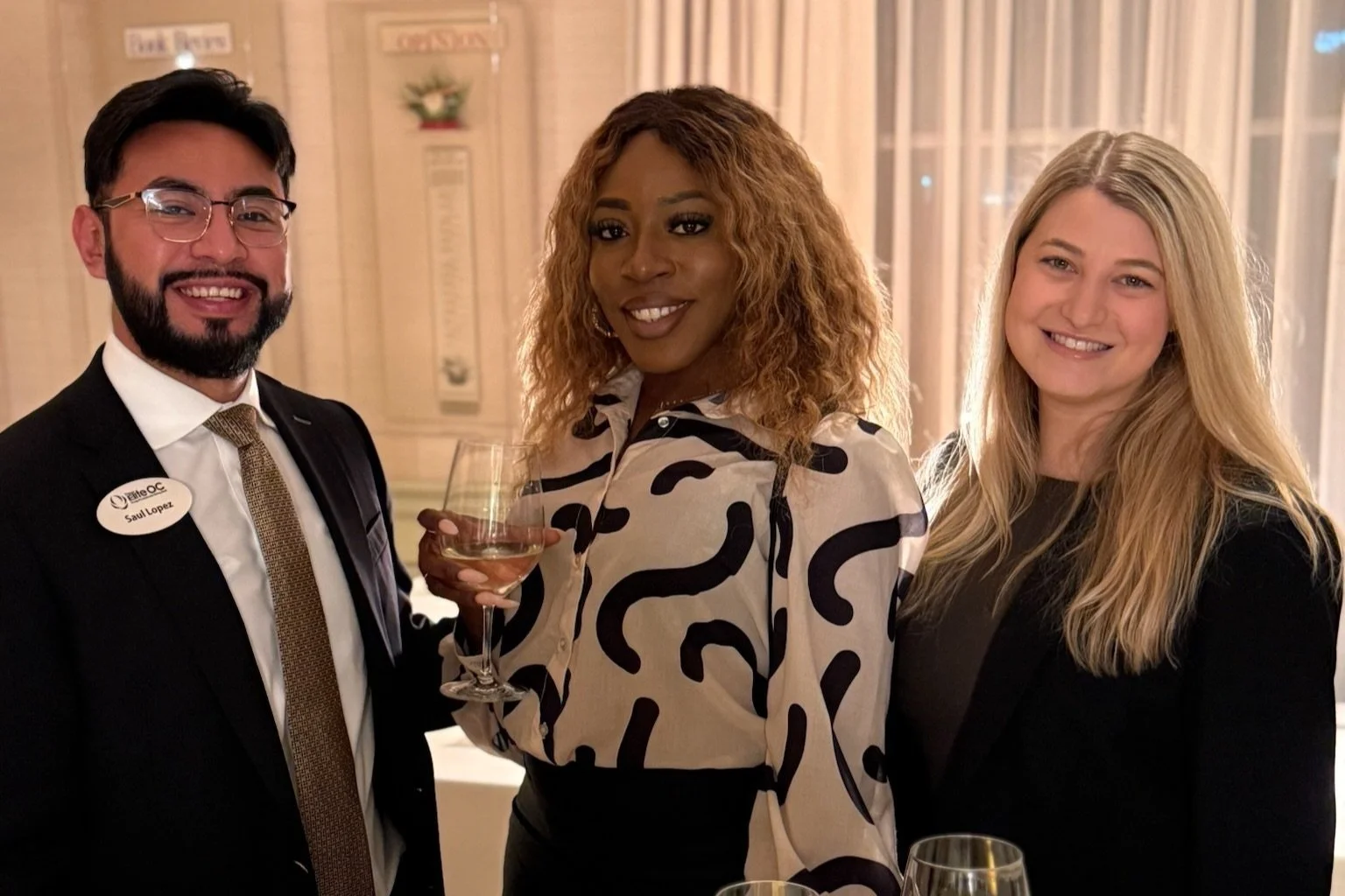 Three people dressed in formal attire at a social event, with two women and one man smiling and holding a wine glass, in an elegant room with drapes and warm lighting.