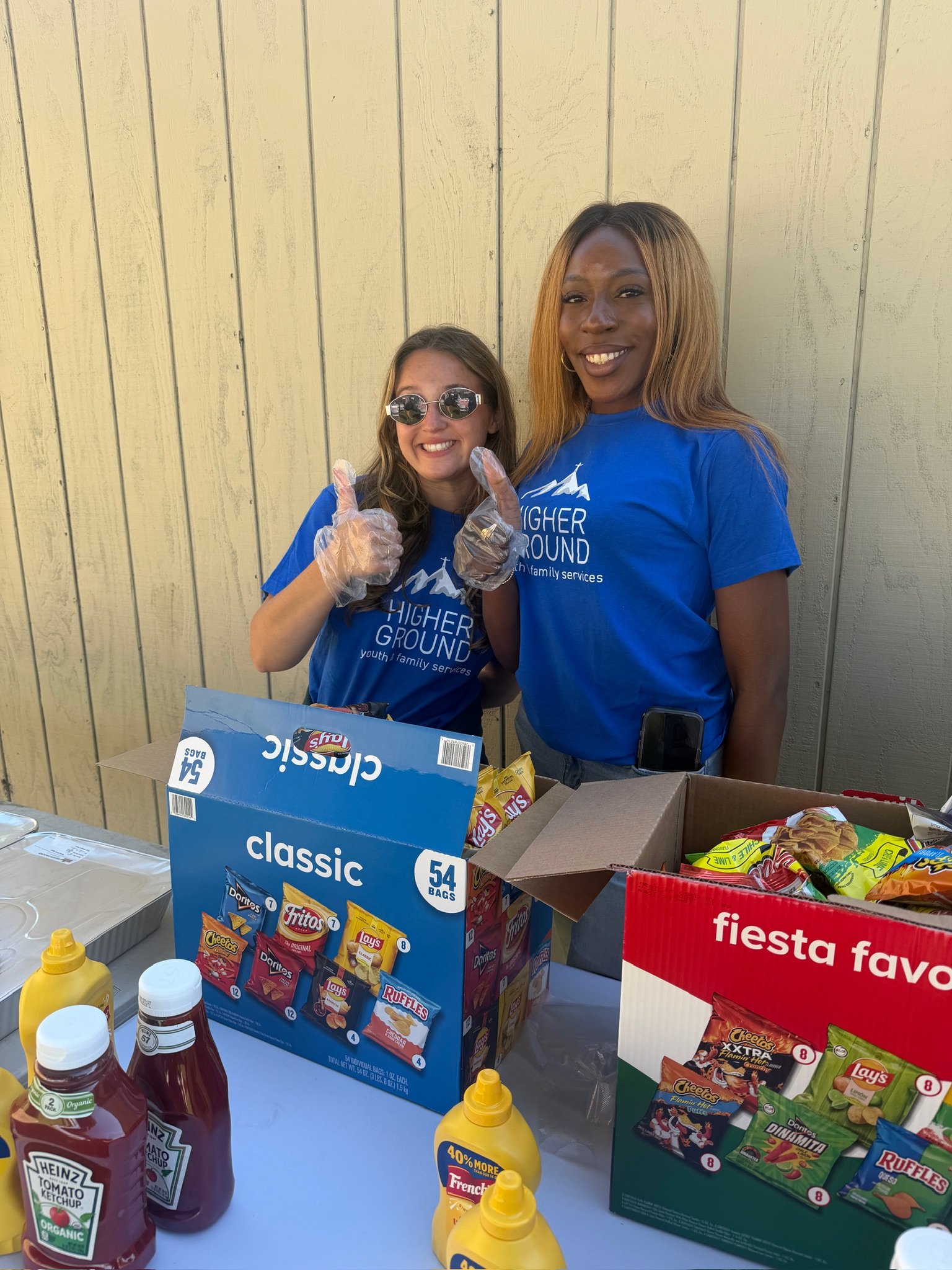 Two women standing behind a table of snacks at an outdoor event. One woman is giving a thumbs-up and the other is smiling. They are wearing blue shirts with a logo that says 'Higher Ground youth & family services.' There are boxes of chips, mustard, ketchup, and other snacks on the table.