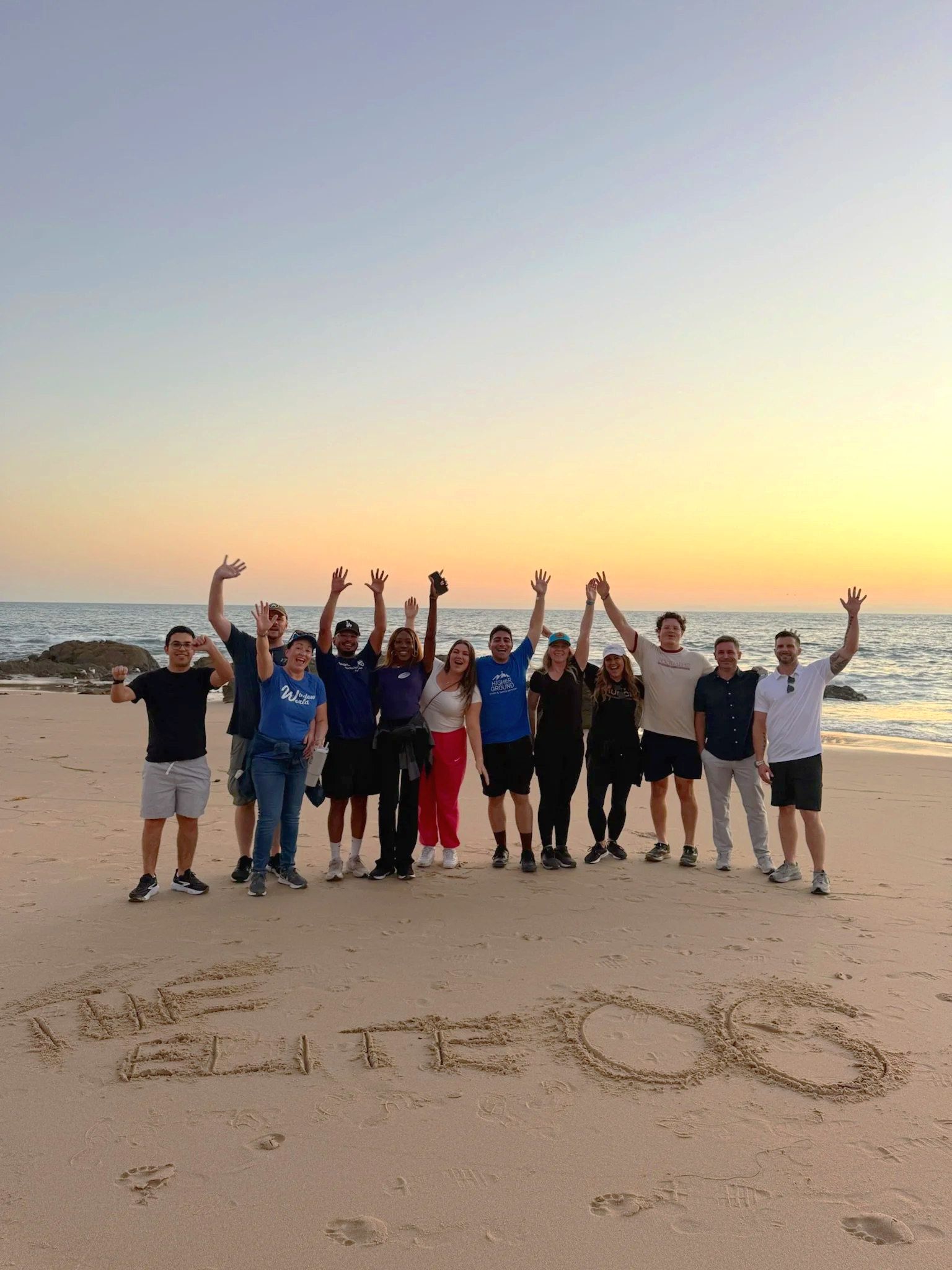 A group of people on a beach during sunset, with some raising their hands and smiling, and 'WE LOVE YOU' written in the sand in the foreground.